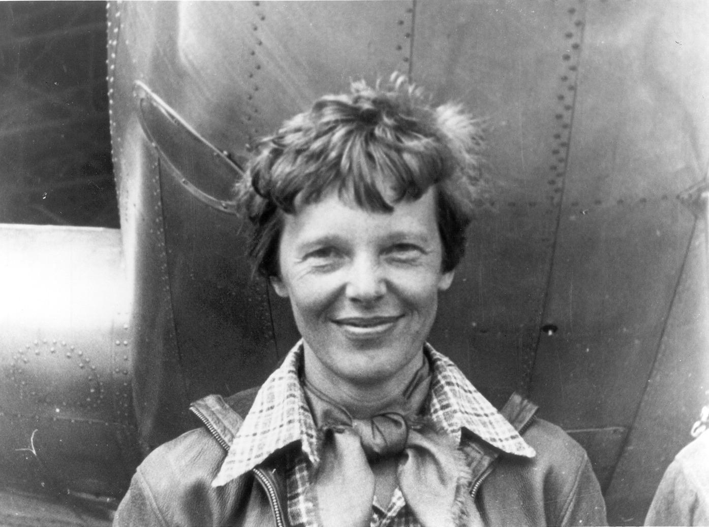 A black-and-white photograph of Amelia Earhart smiling in a leather flight jacket and checked scarf, standing in front of the riveted aluminium fuselage of her Lockheed Electra.