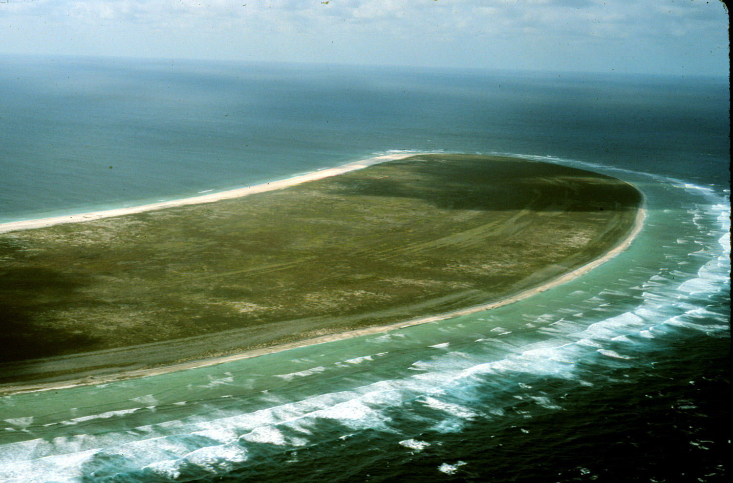 An aerial view of Howland Island — a tiny, flat, teardrop-shaped island of green scrub and pale sand ringed by white surf, set alone in the deep blue of the open Pacific Ocean stretching to the horizon.