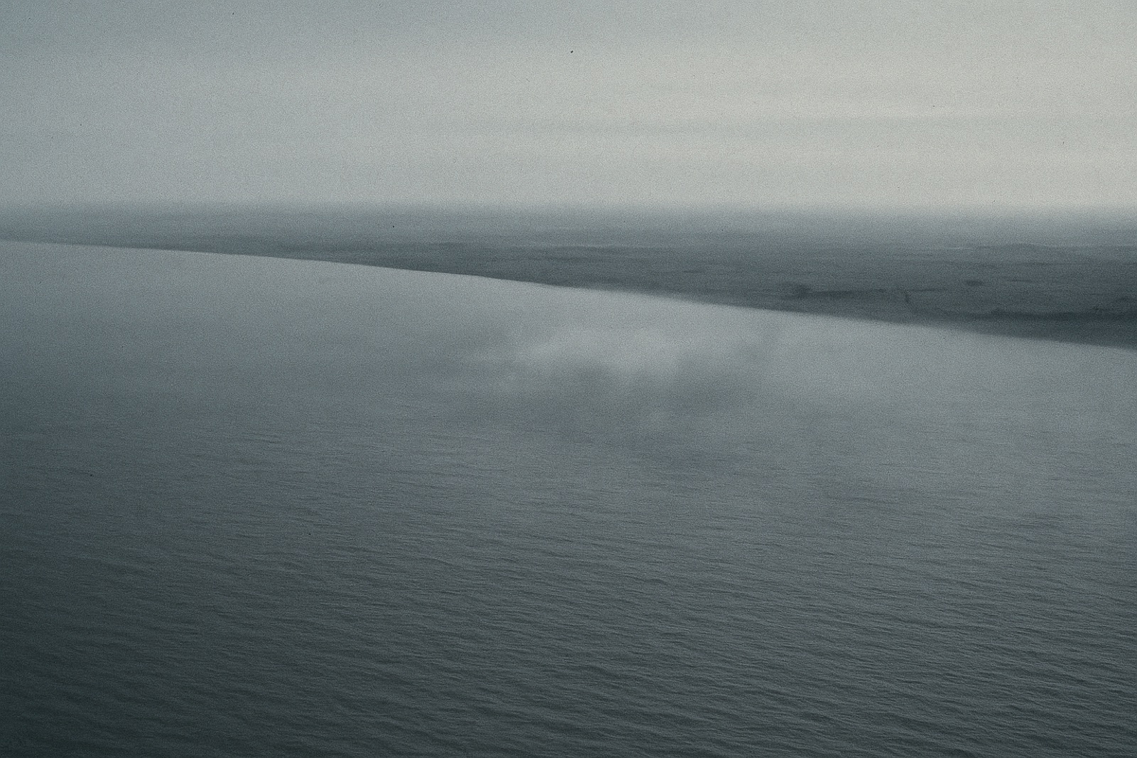 An aerial view at dawn looking down at the Río de la Plata estuary — a flat expanse of brown-grey water meeting a thin coastline in the upper third of the frame, with faint mist on the surface.