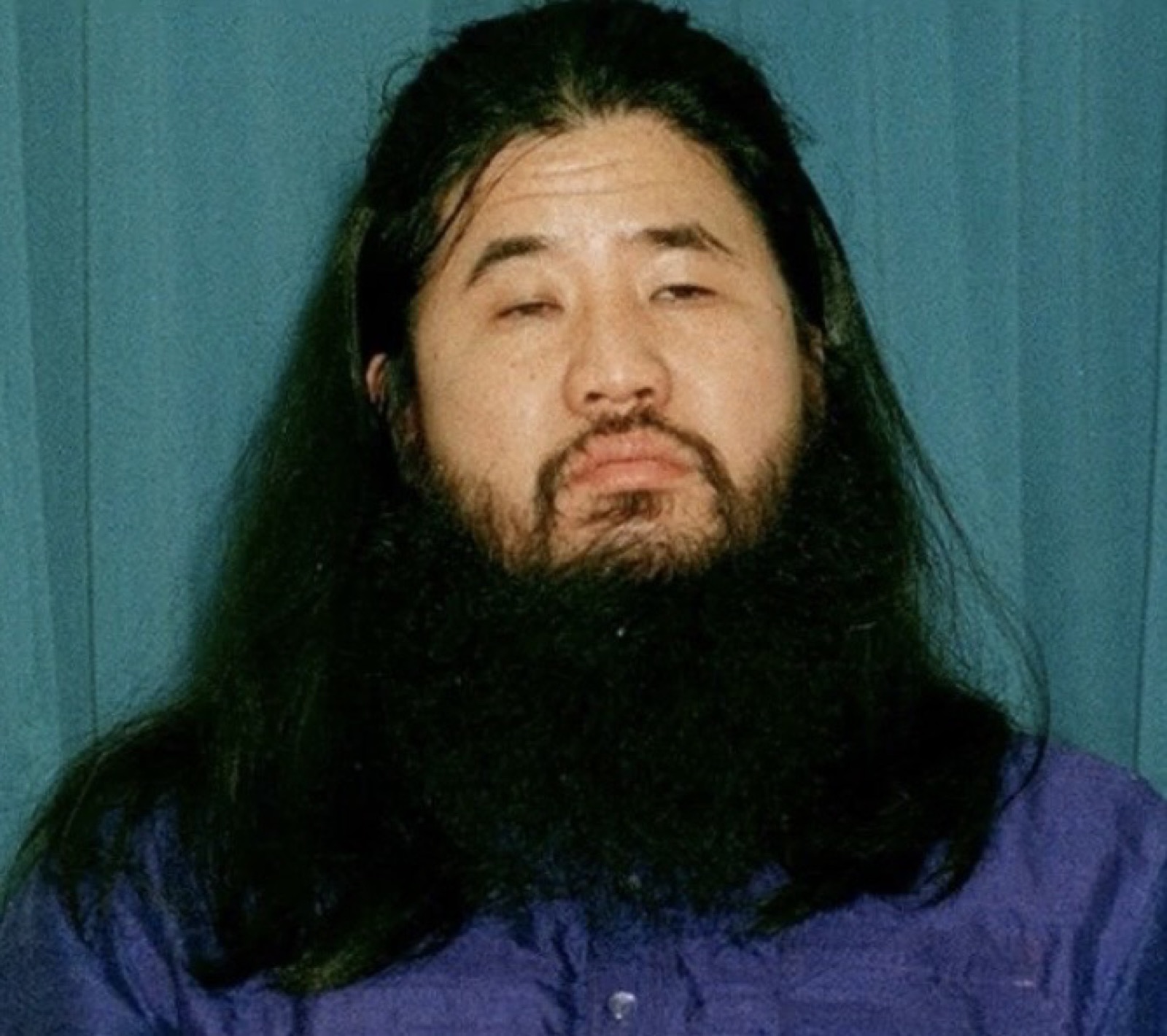 Shoko Asahara photographed seated — bearded, eyes partially closed, wearing dark traditional Japanese robes.