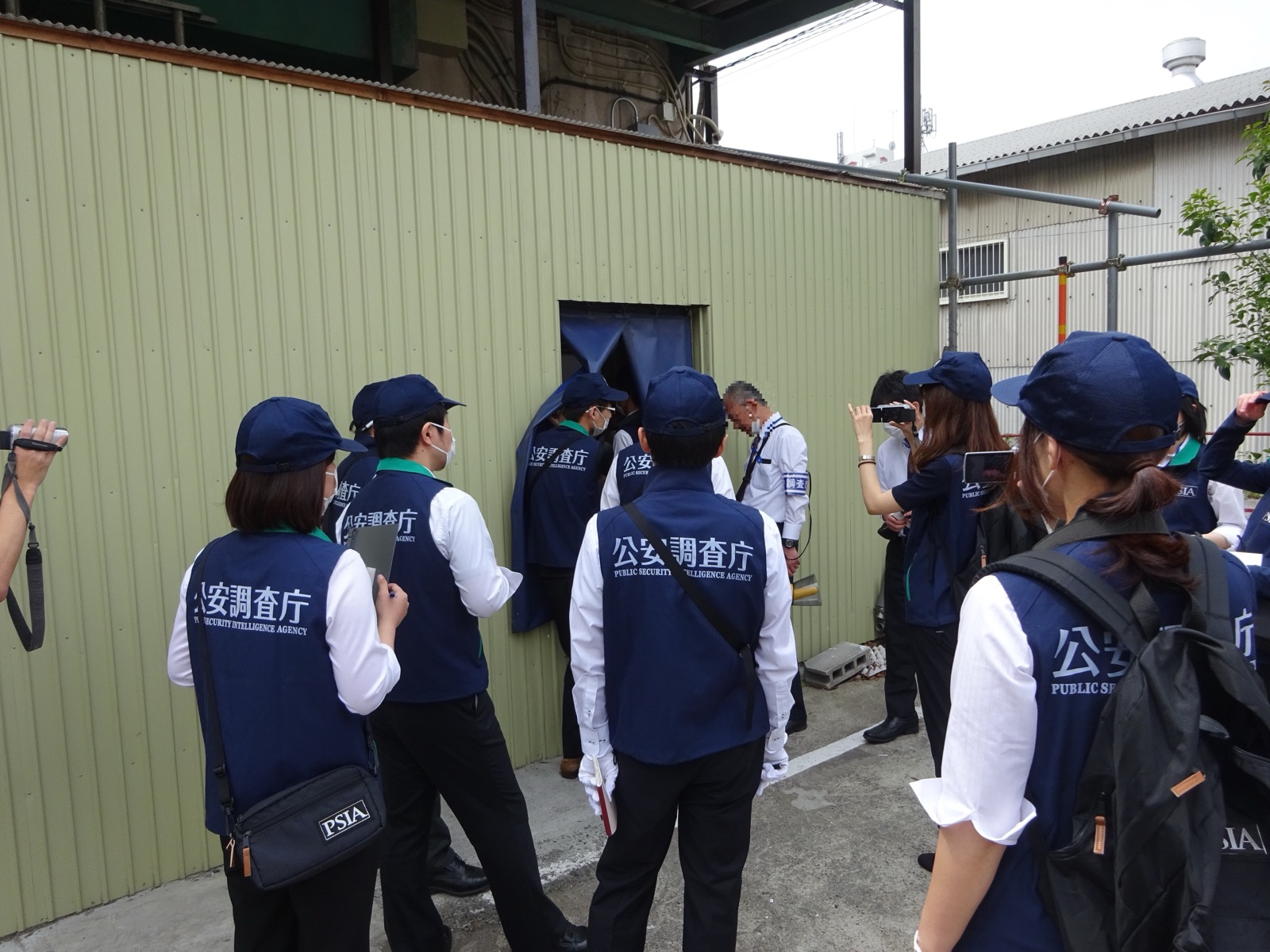 A Japanese Public Security Intelligence Agency officer inspecting a former Aum Shinrikyo facility — uniformed personnel checking a building exterior.