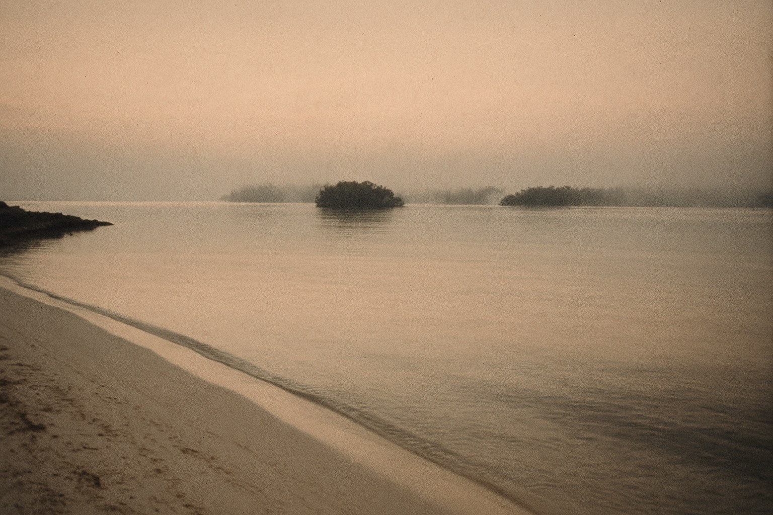 An empty stretch of Cuban Caribbean coastline at first light — white-sand beach in the foreground, calm dark coral-fringed shallow water in middle distance, low scattered mangrove islets on the horizon, overcast sky lit faintly pink-orange from sunrise. No boats, no figures.