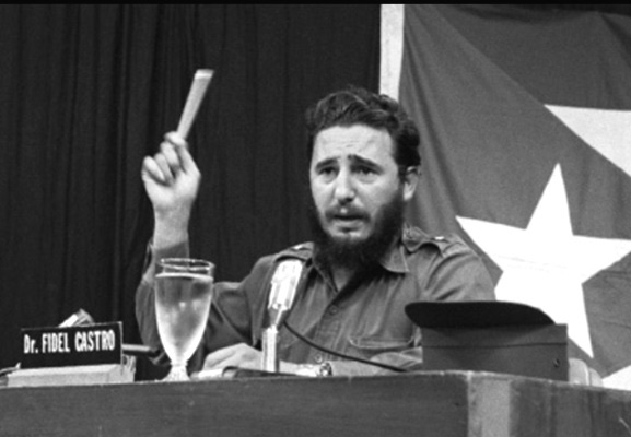 Fidel Castro speaking from a podium with microphones in 1961 — bearded, in olive military uniform, gesturing with his hand mid-speech, surrounded by other officials in military attire.