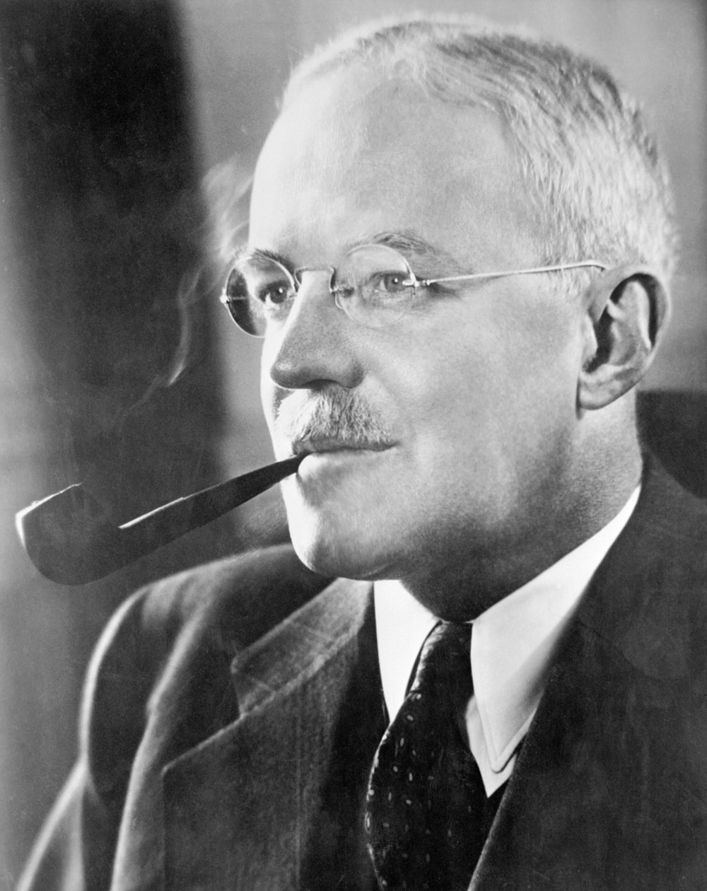 Allen Dulles in his official Central Intelligence Agency portrait — middle-aged man with mustache and round wire-frame glasses, wearing a dark suit, holding a pipe, photographed in formal portrait pose.