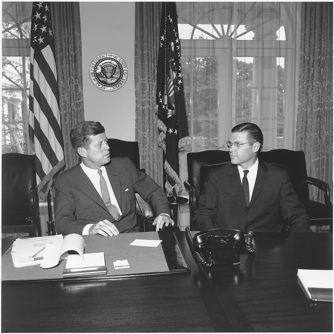 President John F. Kennedy meeting with Secretary of Defense Robert McNamara in the White House Cabinet Room, 1961 — both men seated at the conference table, Kennedy gesturing while speaking, papers and folders on the table between them.
