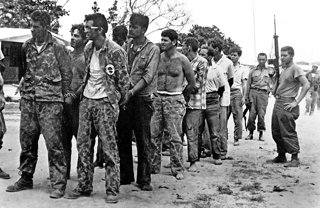 Brigade 2506 personnel as Cuban prisoners after the failed landing, April 1961 — rows of men in disheveled uniforms or civilian clothes, some seated on the ground, others standing, Cuban militia visible in the background.