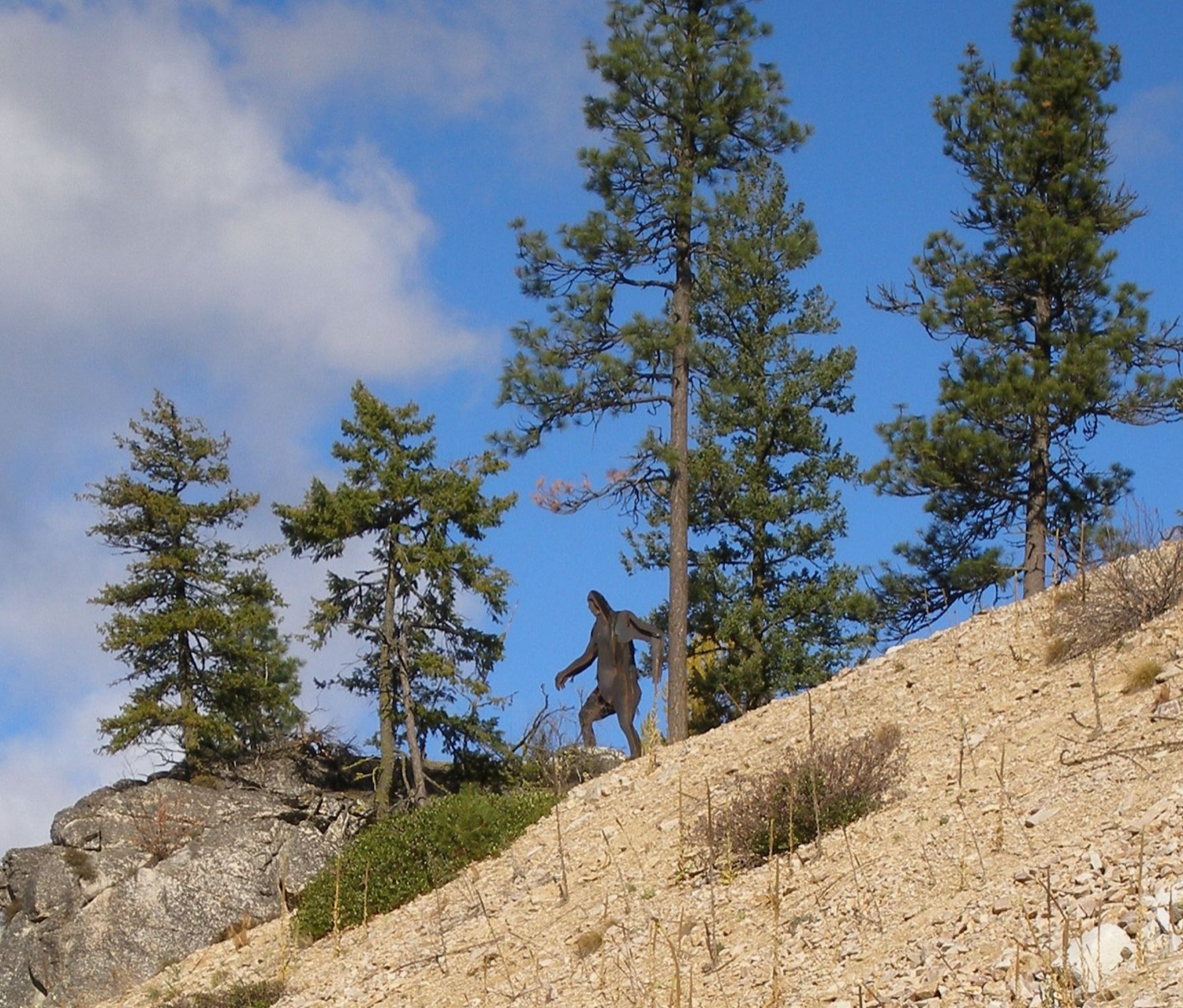 A metal silhouette statue of a striding Sasquatch on a rocky, pine-dotted hillside against a blue sky, a roadside attraction.