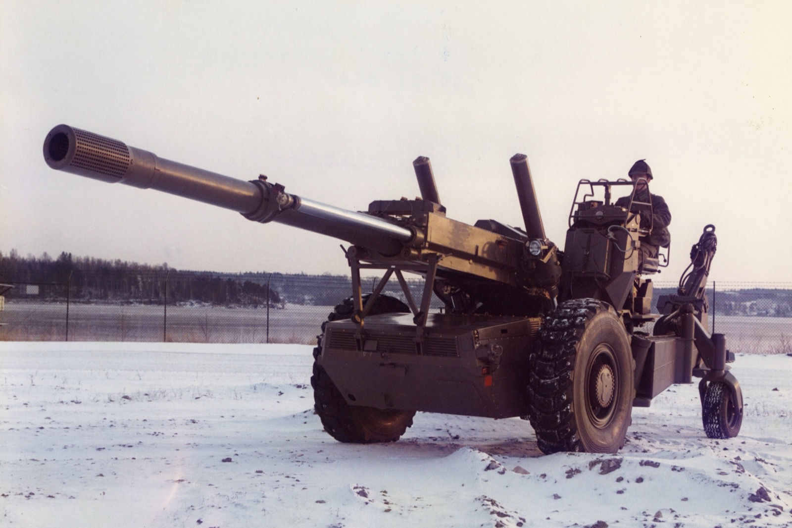 A Bofors FH-77 field howitzer, a large towed artillery piece with a long barrel, standing on a snowy field under a pale sky, a soldier beside it.