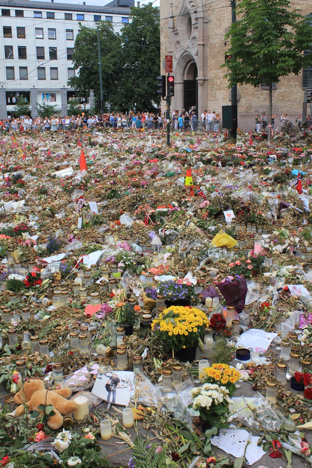 A vast sea of flowers, candles, and tributes covering the plaza in front of Oslo Cathedral in the days after the 22 July 2011 attacks, with people gathered near the cathedral in the background.
