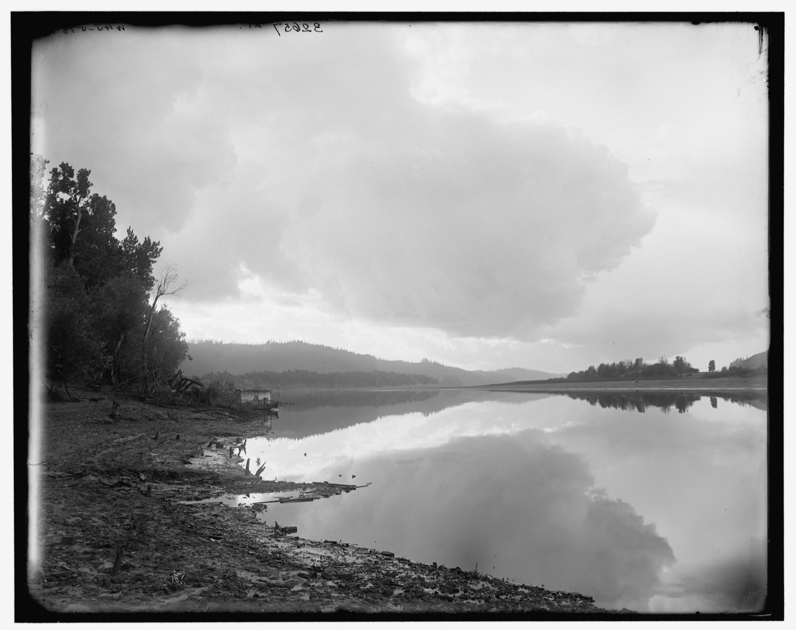 A calm stretch of the Columbia River near Vancouver, Washington, in an old black-and-white photograph — still water reflecting a clouded sky, a wooded shoreline and a low beach in the foreground, forested hills receding into haze.