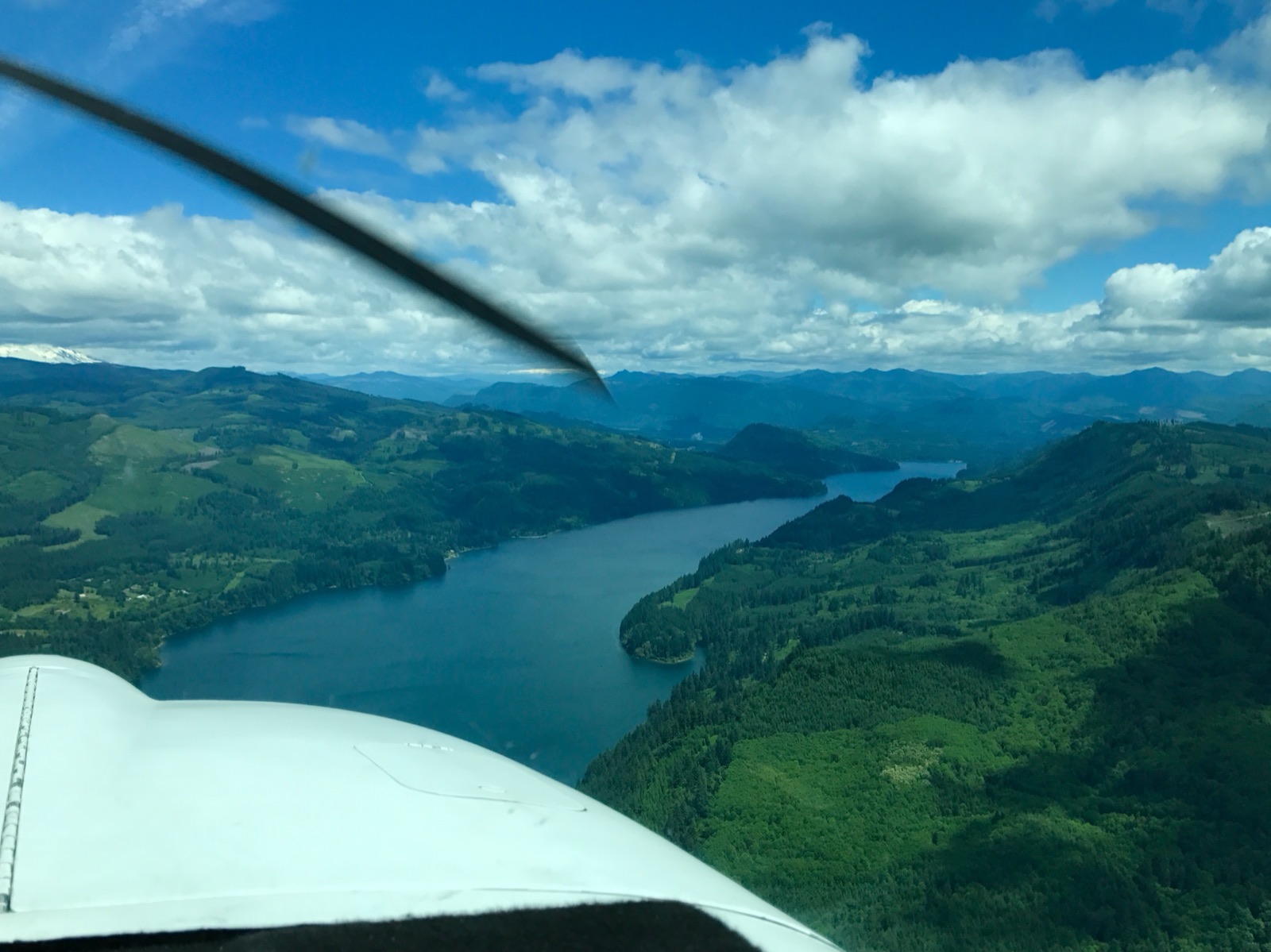 An aerial view of Lake Merwin in southwestern Washington — a long, narrow reservoir of dark green water winding between steep, densely forested ridges under a partly cloudy sky.