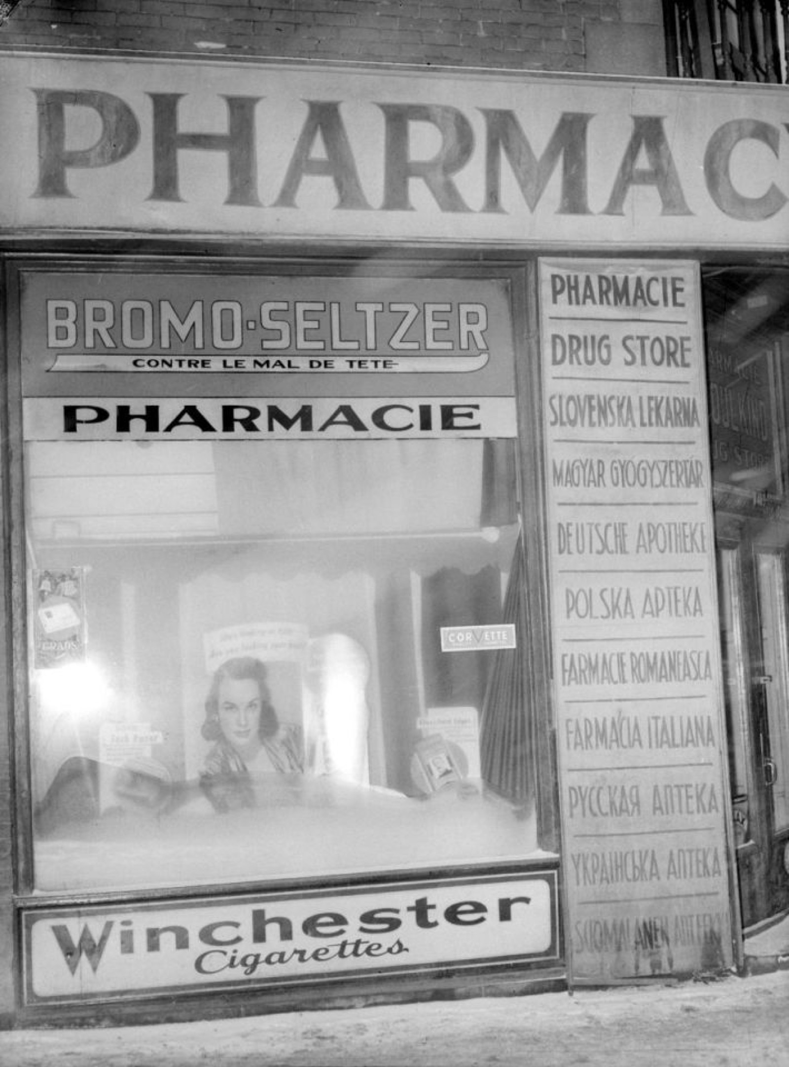 A black-and-white photograph of the interior of a mid-twentieth-century drugstore, with shelves of bottles and a pharmacy counter.