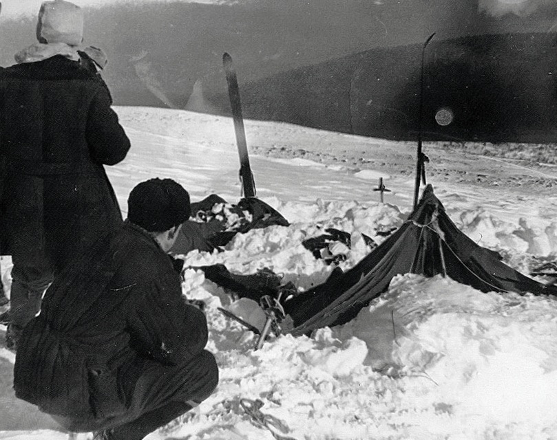 A photograph from the 1959 Soviet search operation — investigators standing near the collapsed Dyatlov tent on the snow slope of Kholat Syakhl, the tent half-buried in snow, slashes visible in the canvas.