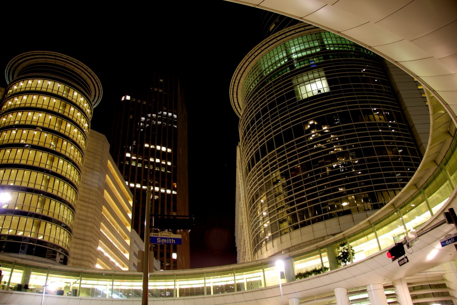 The Enron office complex in downtown Houston at night — two tall office towers, one cylindrical and glass-walled, connected by a curved skybridge over Smith Street, lit up against a dark sky.