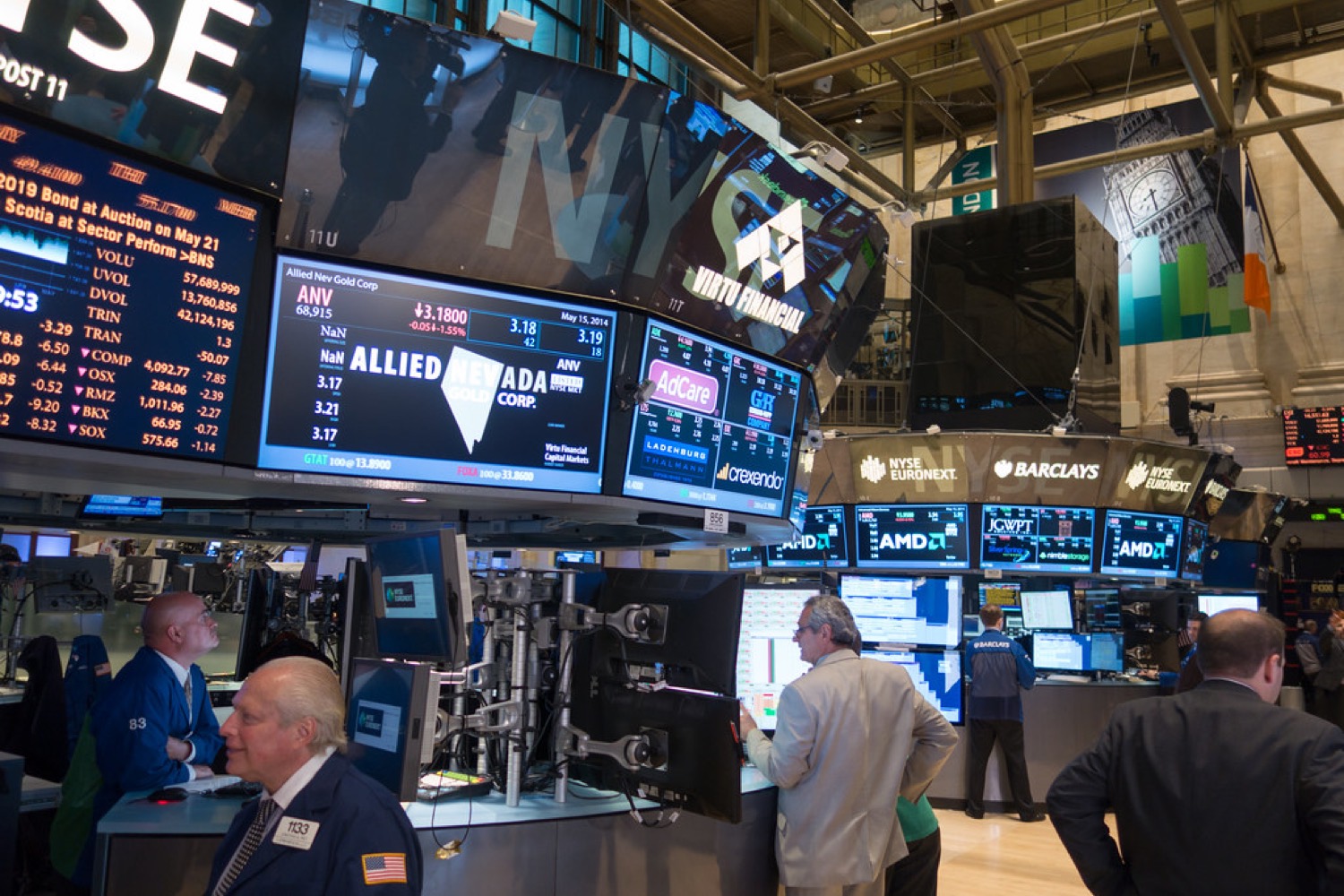 A bustling stock-exchange trading floor with traders at workstations surrounded by screens.