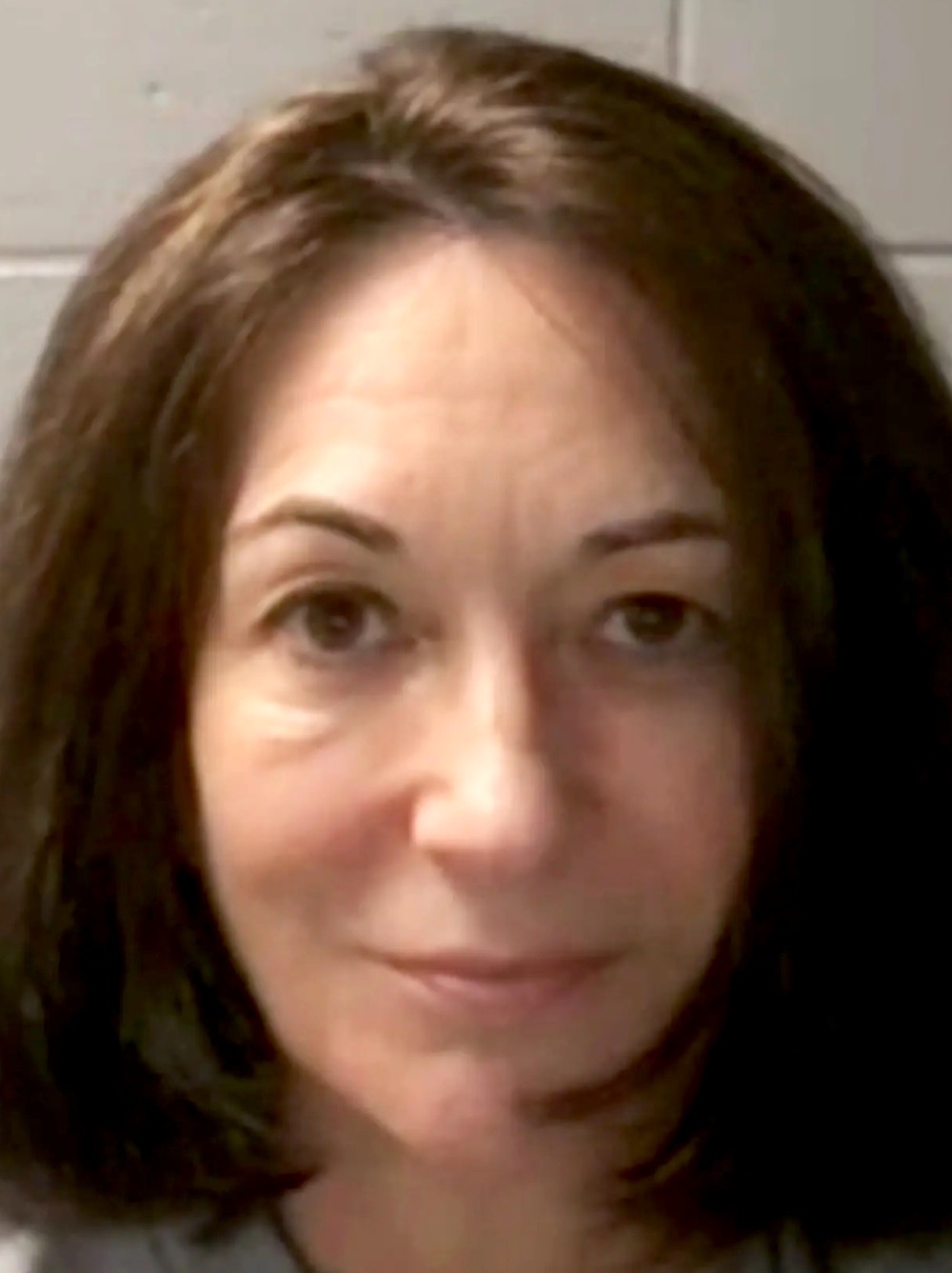 Ghislaine Maxwell in her 2020 federal mug shot — shoulder-length brown hair, neutral expression, photographed against a beige institutional background.