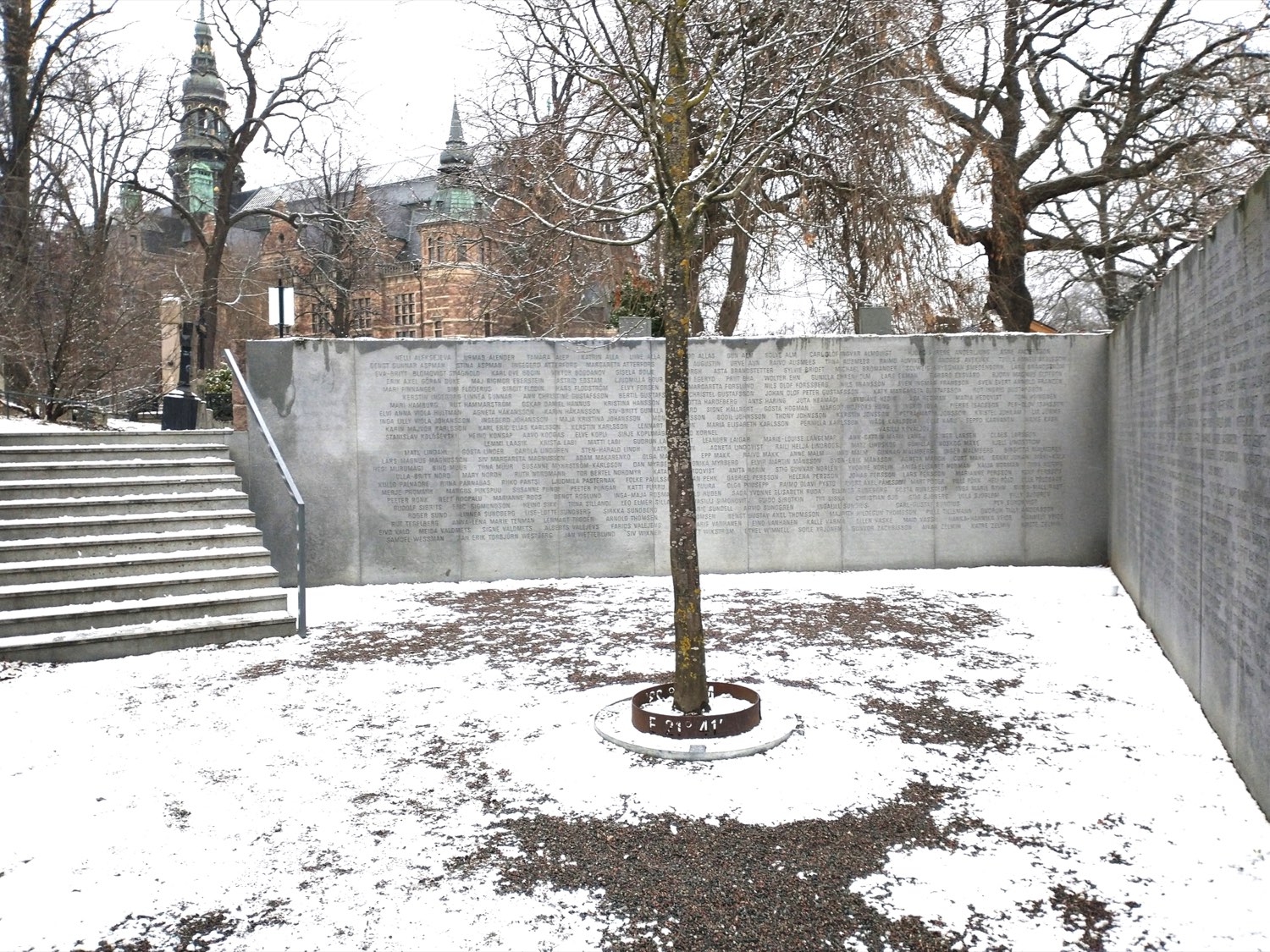 The Estonia memorial in Stockholm — curved stone walls engraved with the names of the dead enclosing a small courtyard with a single bare tree at its centre, lightly dusted with snow, the Nordic Museum visible behind.