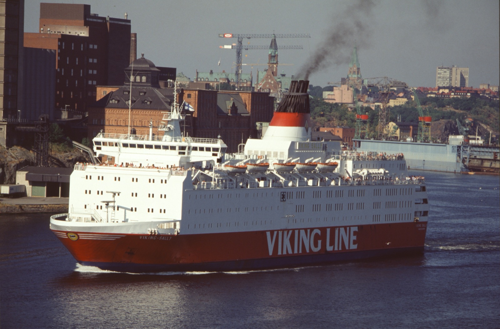 A large white and red passenger ferry, the Viking Sally, sailing in a harbour with a city skyline behind, the words 'VIKING LINE' on her red hull.