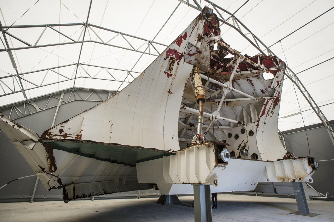 The recovered bow visor of MS Estonia, a large white metal clamshell structure with torn and rusted edges, displayed on supports inside a covered storage hall.