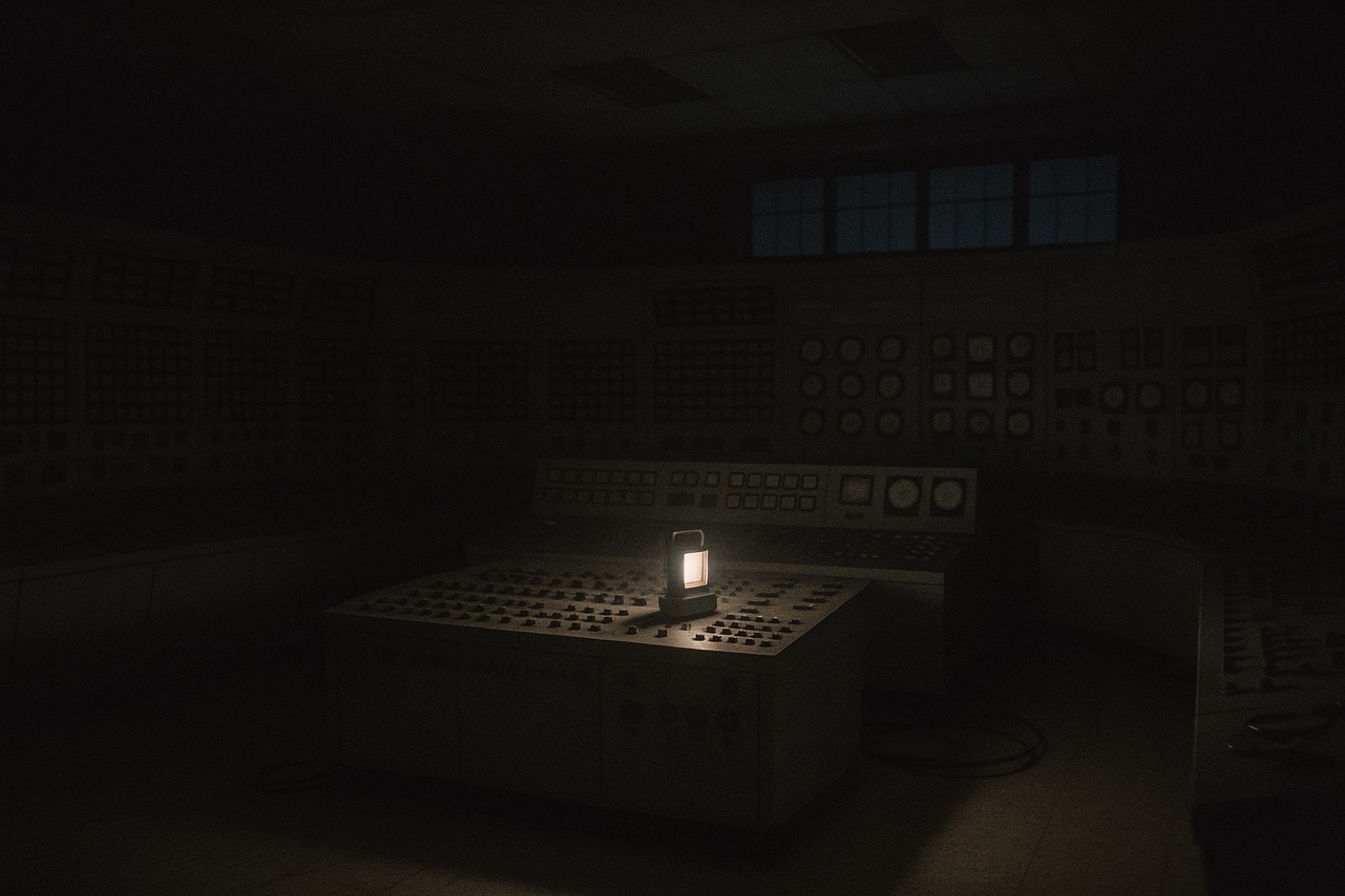 The main control room of an older boiling-water nuclear power station in near-total darkness during a total loss of power — long curved analog panels covered in hundreds of small indicator lamps, rotary switches, dials and chart recorders, every light dead, lit only by a single battery emergency lantern on the console and a faint blue-grey glow from the windows. The room is empty. No people.