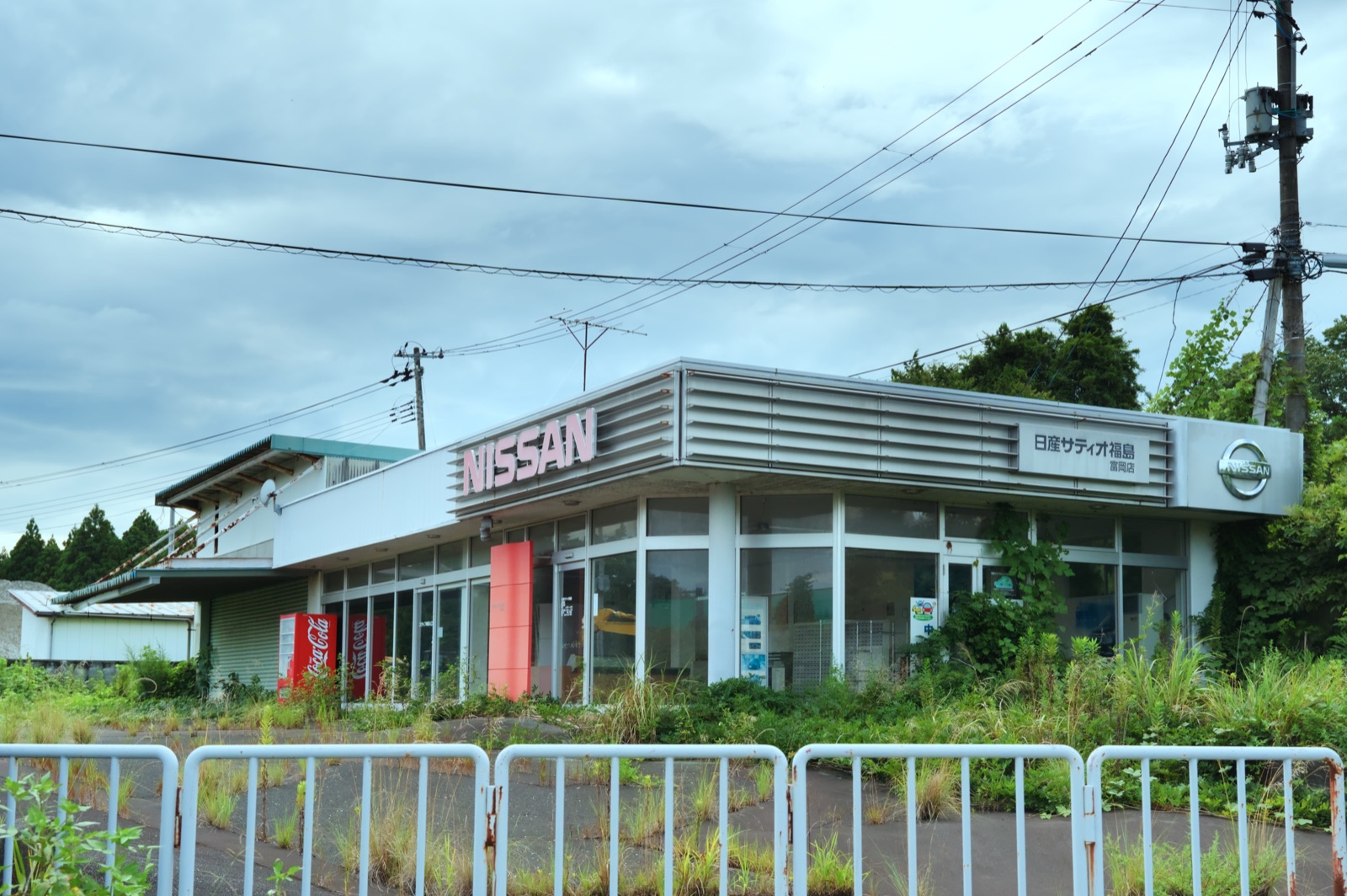 An abandoned Nissan car dealership inside the Fukushima evacuation zone — a single-storey glass-fronted showroom with a faded NISSAN sign, weeds and tall grass growing up through the forecourt, rusting vending machines, and a low barrier fence across the front, under an overcast sky. No people.