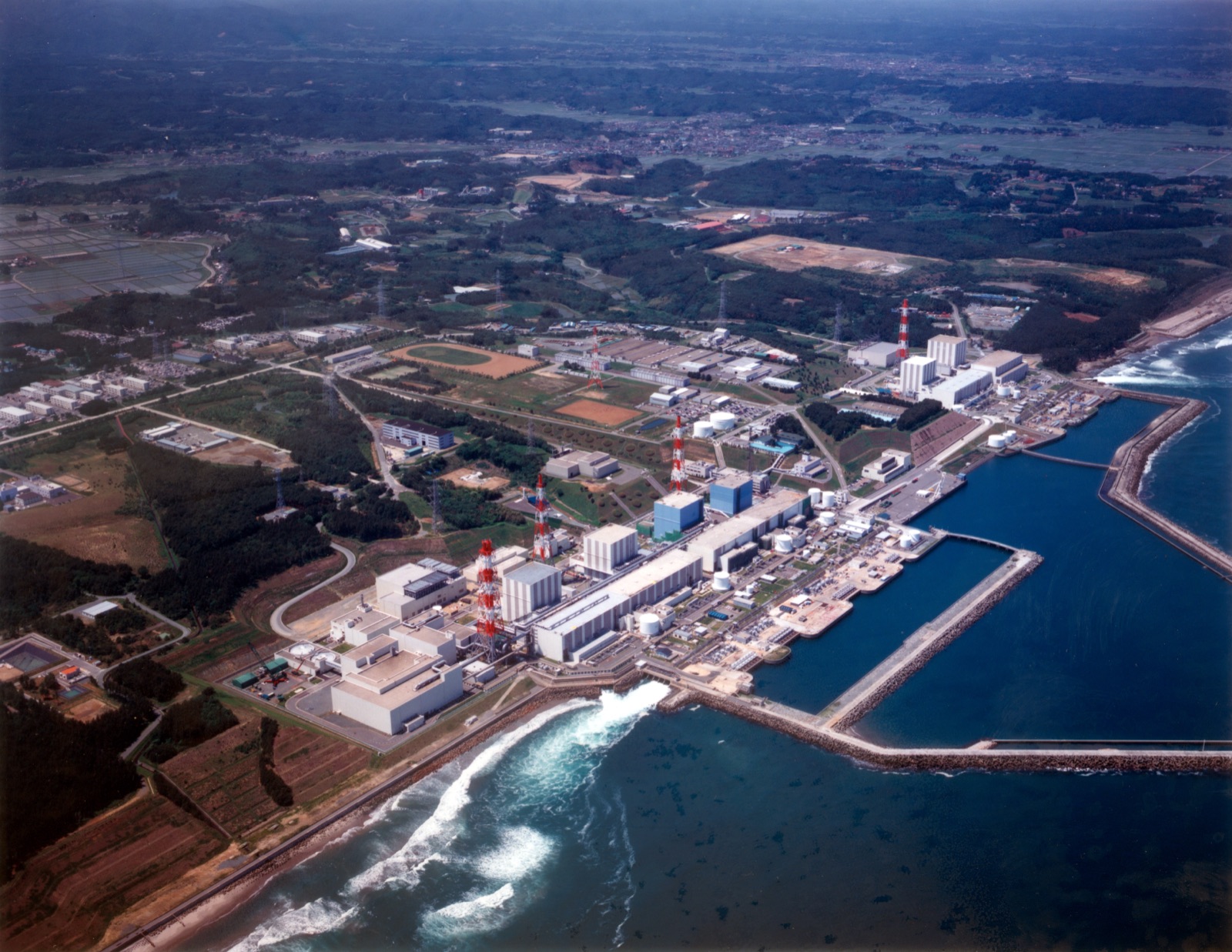 An aerial photograph of the Fukushima Daiichi Nuclear Power Plant before the disaster — a row of reactor and turbine buildings with tall red-and-white exhaust stacks strung along a low coastal shelf, the breakwaters of the plant harbour reaching into the Pacific on the right, farmland and wooded hills inland on the left.
