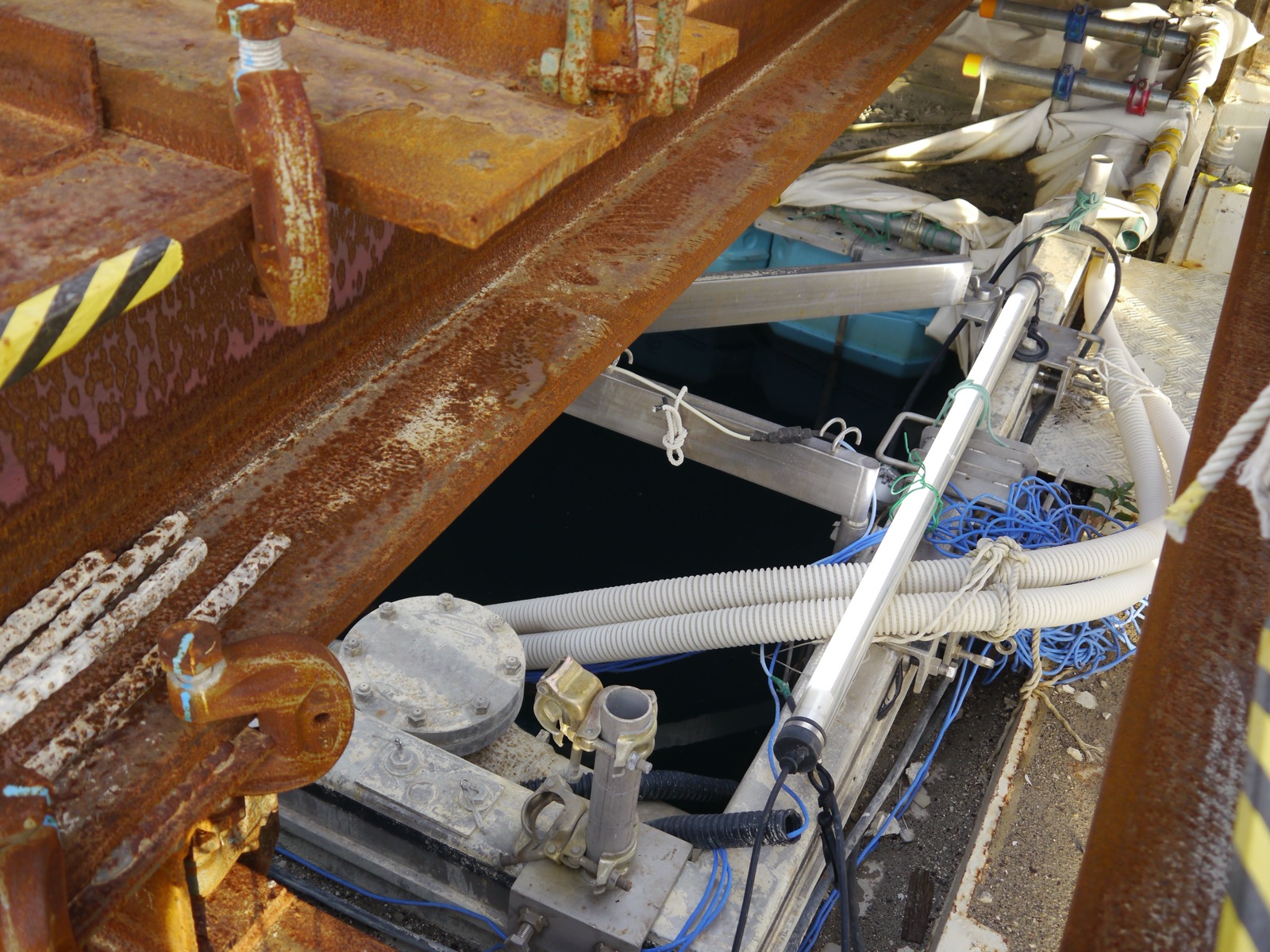 A close-up of improvised recovery equipment at a Fukushima Daiichi reactor — rusted steel beams, a tangle of grey hoses and blue cables, valves and fittings, rigged over the dark water of a spent fuel pool during an inspection.