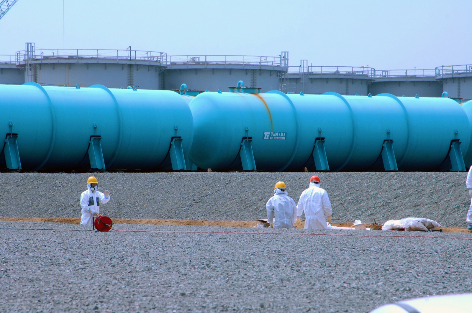 Rows of very large pale-blue cylindrical water storage tanks at the Fukushima Daiichi site, lying on a gravel embankment, with a few workers in white protective suits and hard hats standing small in the foreground for scale.