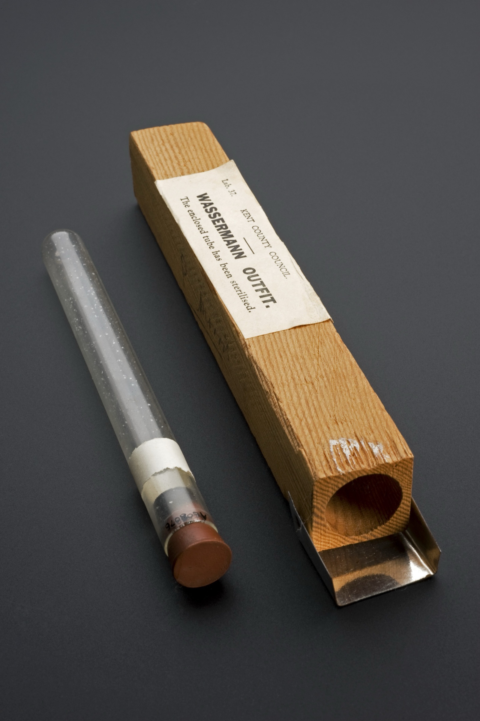 A glass specimen bottle of the kind used to collect blood for a syphilis test, with a paper label, photographed against a plain background.