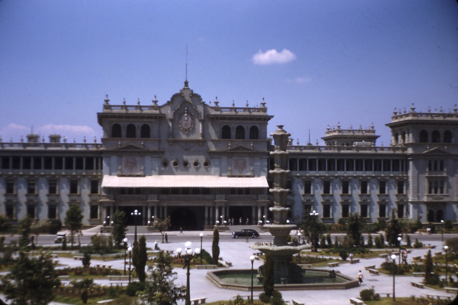 A colour photograph from 1948 of the National Palace of Guatemala in Guatemala City — a large pale green-grey palace behind a plaza with a tiered fountain, gardens, and a few vintage cars under a blue sky.