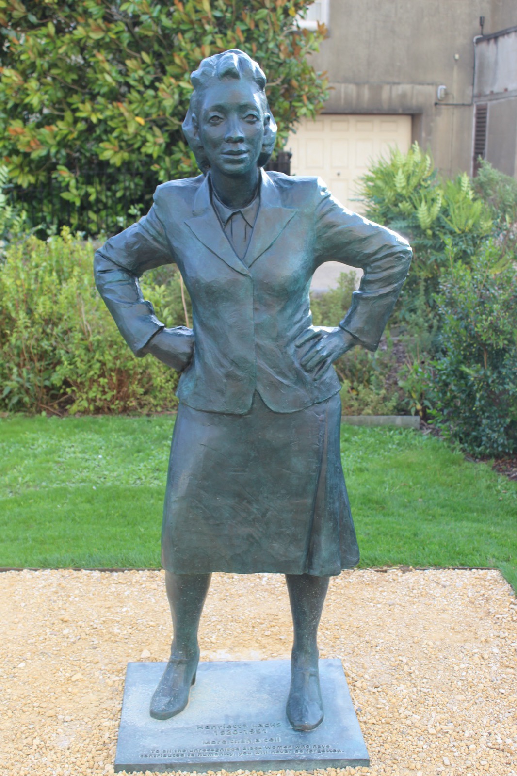 A bronze statue of Henrietta Lacks standing with her hands on her hips, wearing a 1950s skirt suit, on a plinth in a garden — the EVE statue in Bristol, England.