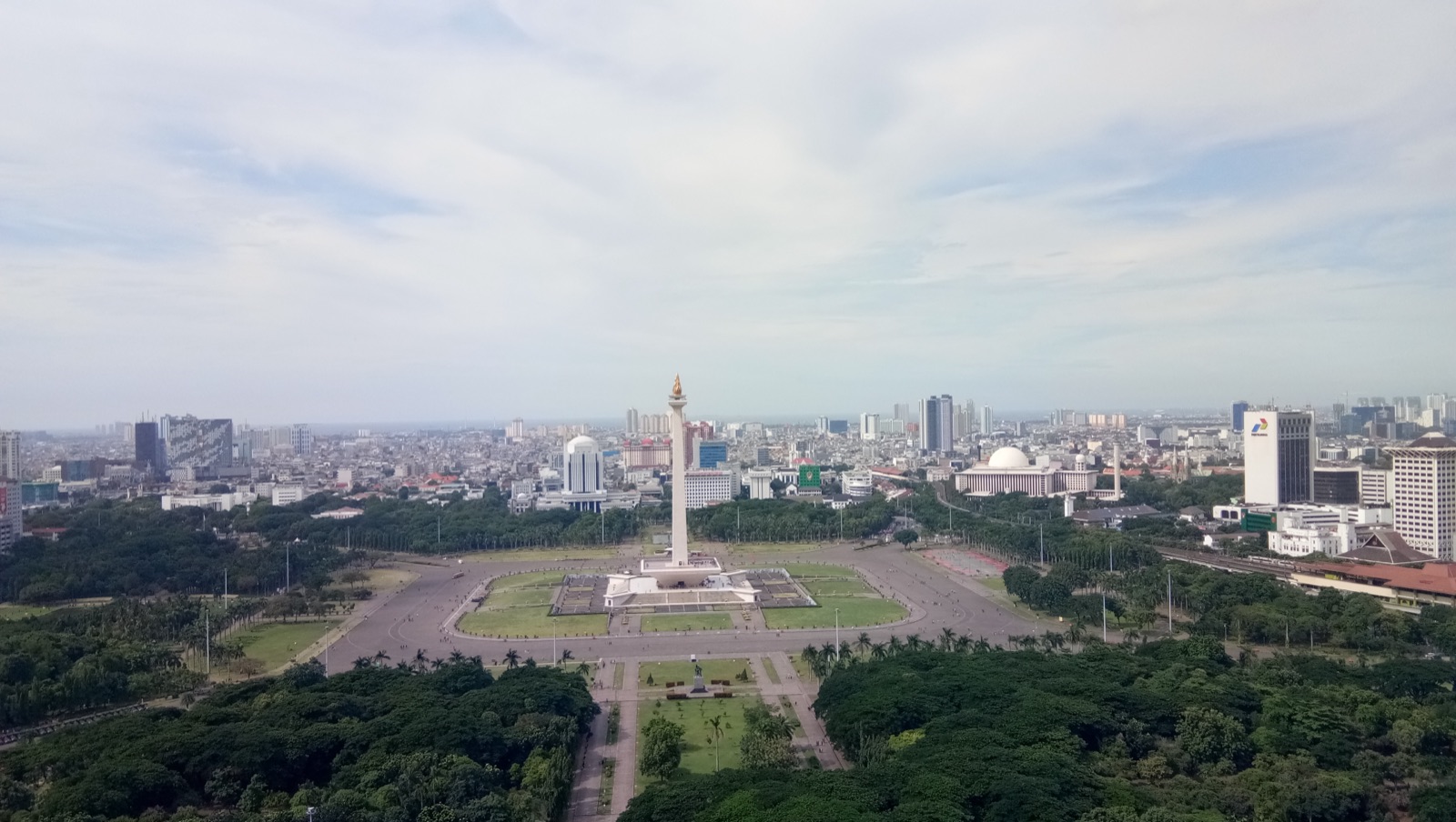 The National Monument (Monas) in central Jakarta, photographed in daylight — a tall obelisk topped with a gold-leaf flame, surrounded by a wide public plaza.