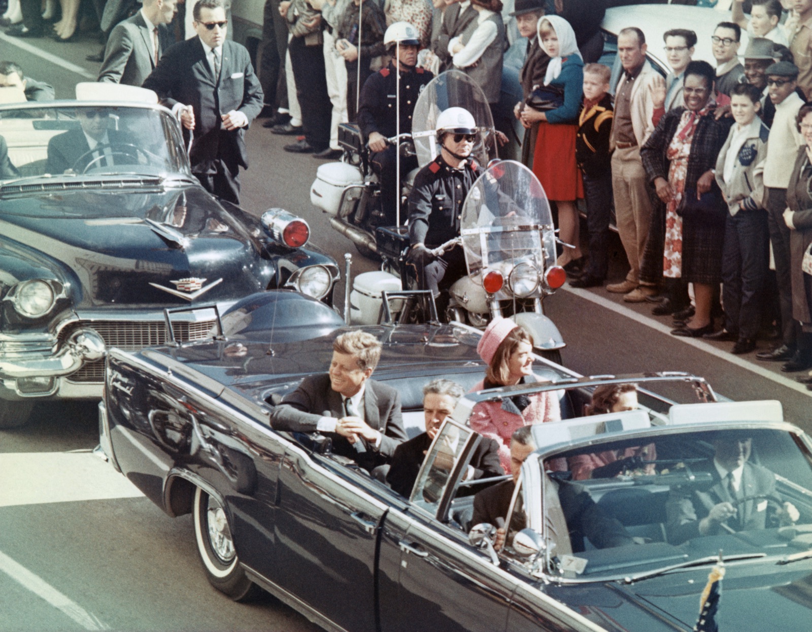 The Kennedy motorcade in Dallas — the presidential limousine with President Kennedy and Mrs. Kennedy in the back seat, Texas Governor John Connally and his wife in the jump seats, Secret Service agents visible.