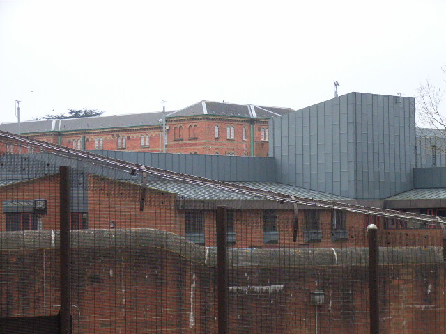 The exterior entrance to Broadmoor Hospital — a tall red-brick Victorian wall enclosing the high-security psychiatric hospital, with the gatehouse and tree line visible.