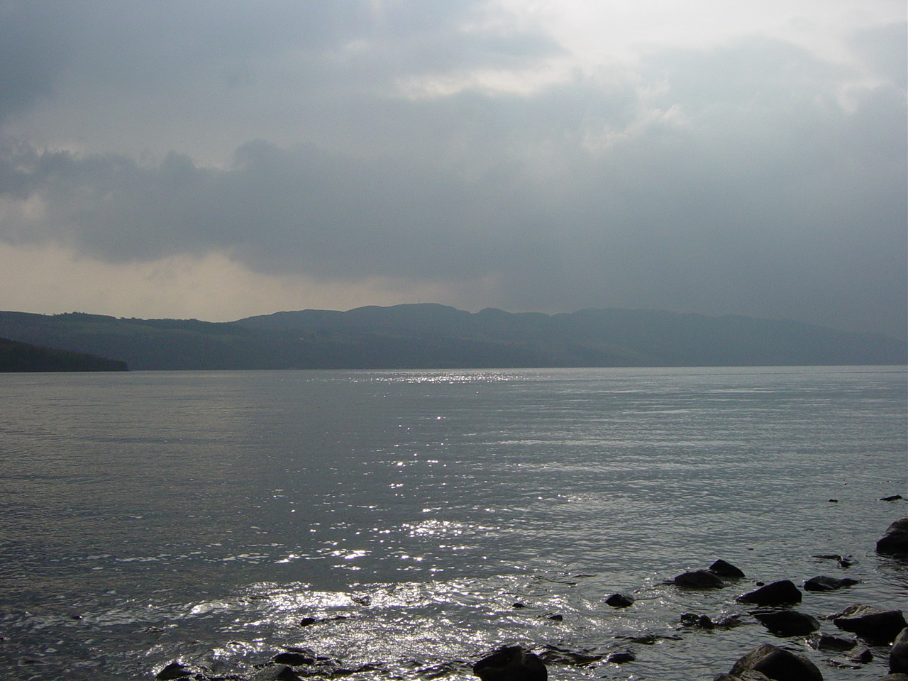 Loch Ness under a low mist — the calm, dark surface of the water fading into grey fog, with the dim outline of hills behind, and no clear horizon.