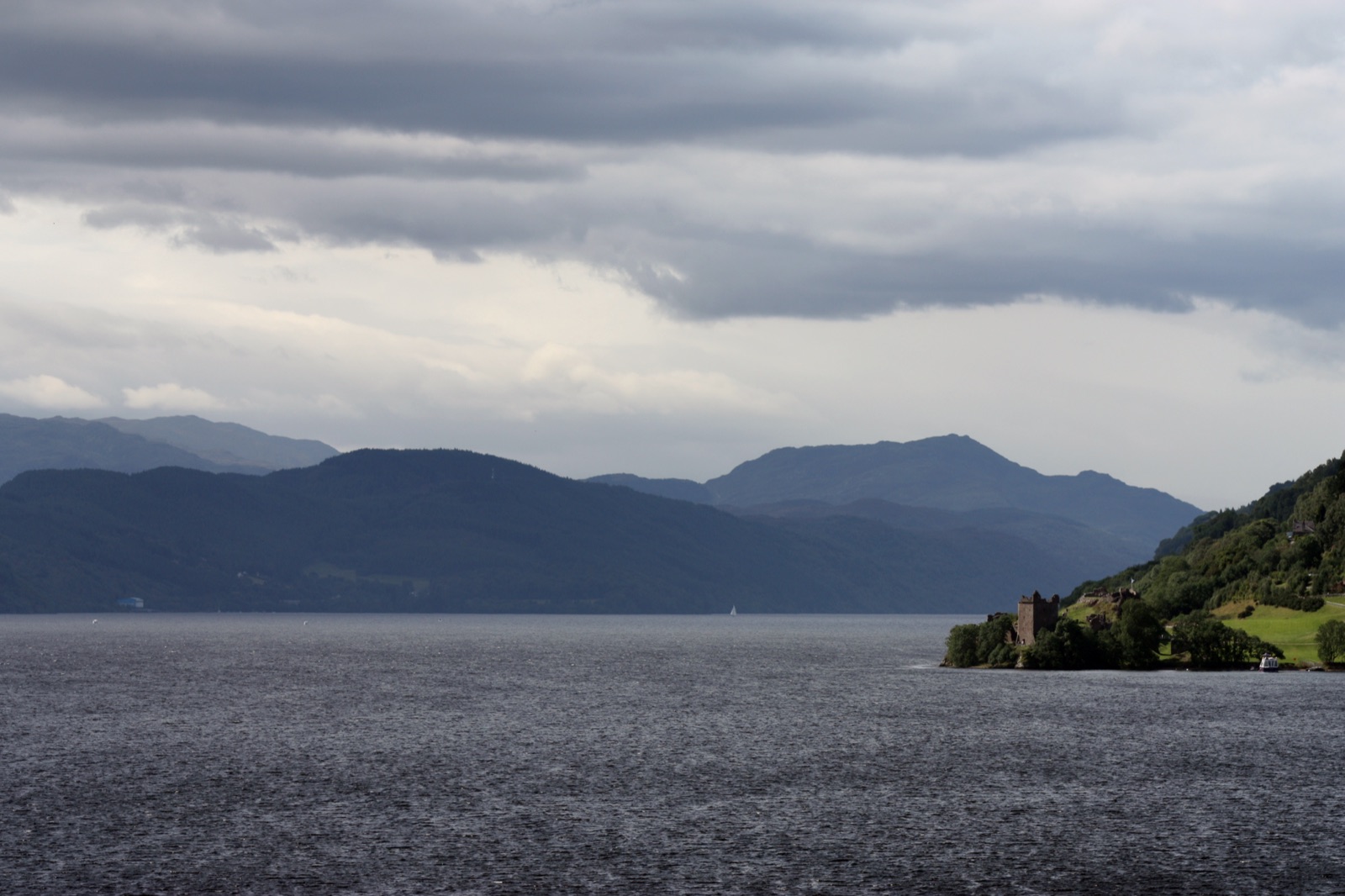A wide view of Loch Ness under heavy grey cloud — a long expanse of dark, slightly rippled water flanked by steep wooded hills, with the small ruin of Urquhart Castle on a promontory on the right shore.