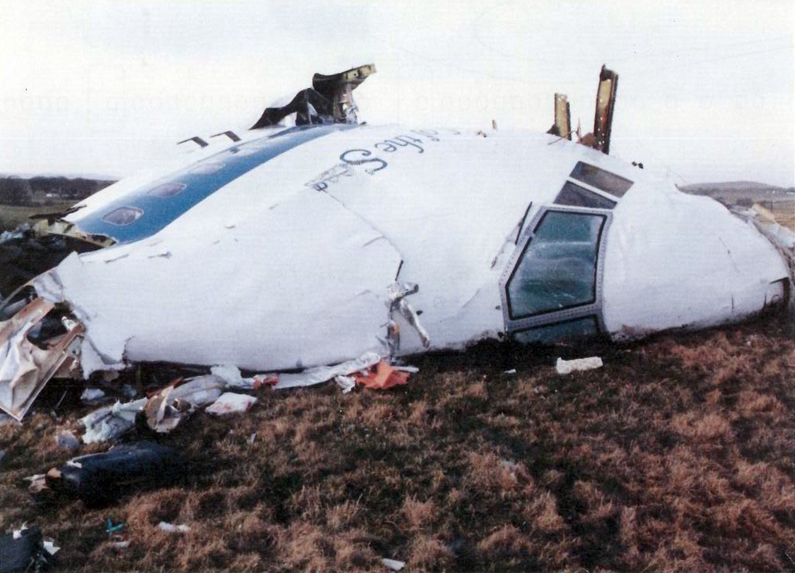 The forward fuselage and cockpit section of Pan Am Flight 103 in a field at Tundergarth Mains near Lockerbie, December 22, 1988 — the curved nose of a Boeing 747 lying on its left side in pasture, recovery vehicles and investigators visible in the background.