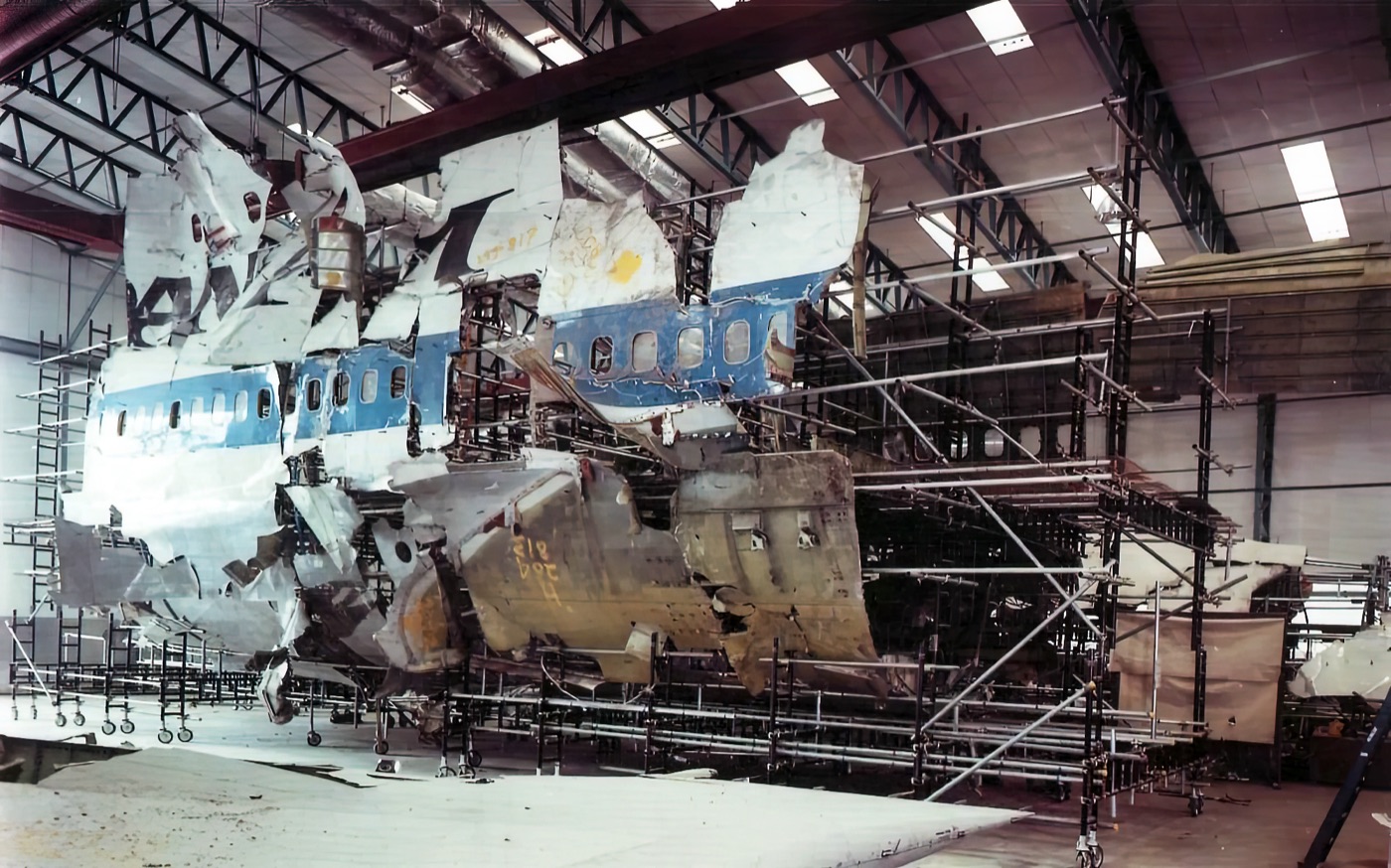 A partial reconstruction of the Pan Am 103 aircraft skin at the AAIB Farnborough facility — visible fuselage panels assembled on a metal frame to map the explosive damage pattern, the reconstructed structure photographed in an indoor hangar.