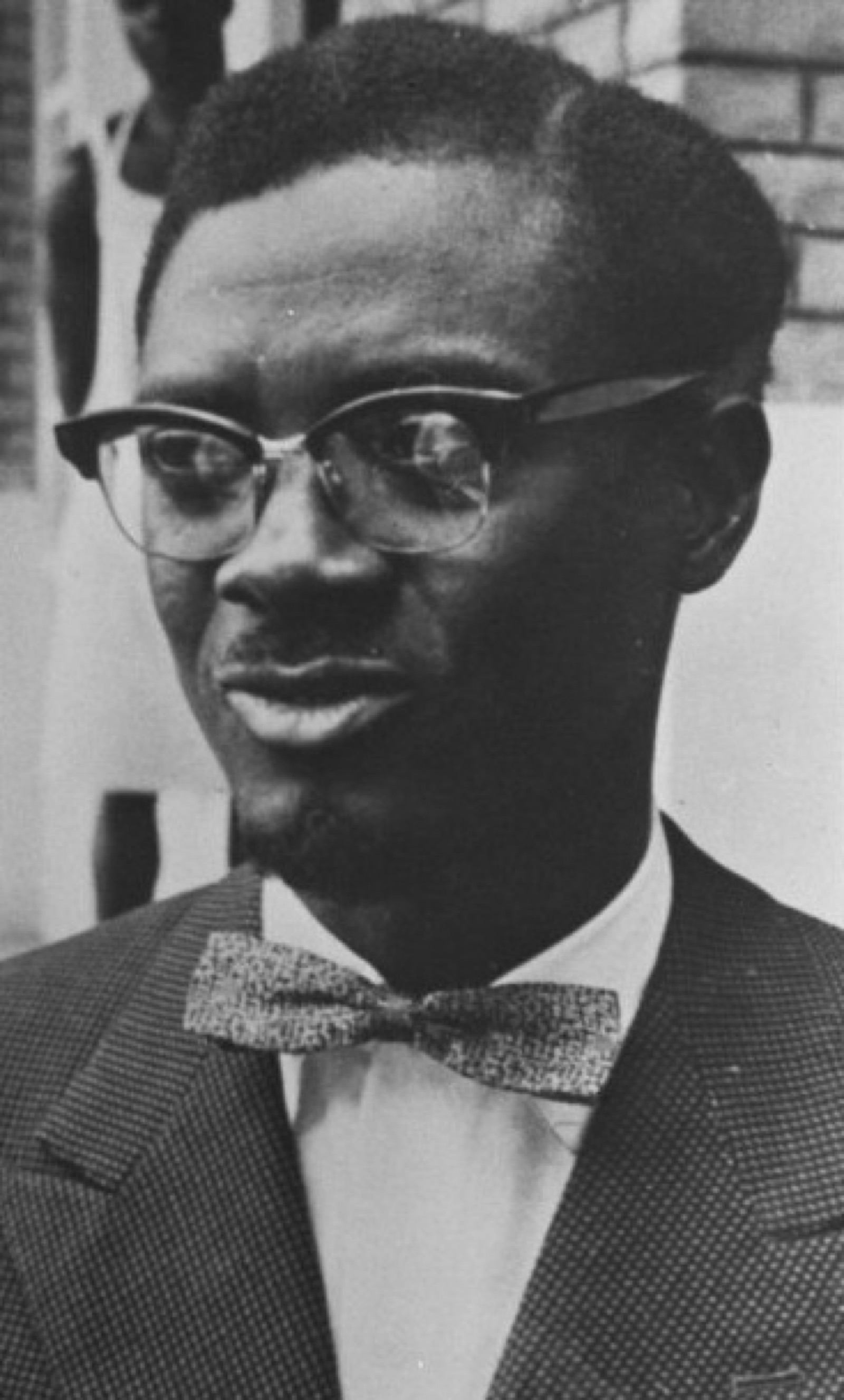Patrice Lumumba photographed in 1960 — wearing a dark suit and tie, looking off-camera with a serious expression.