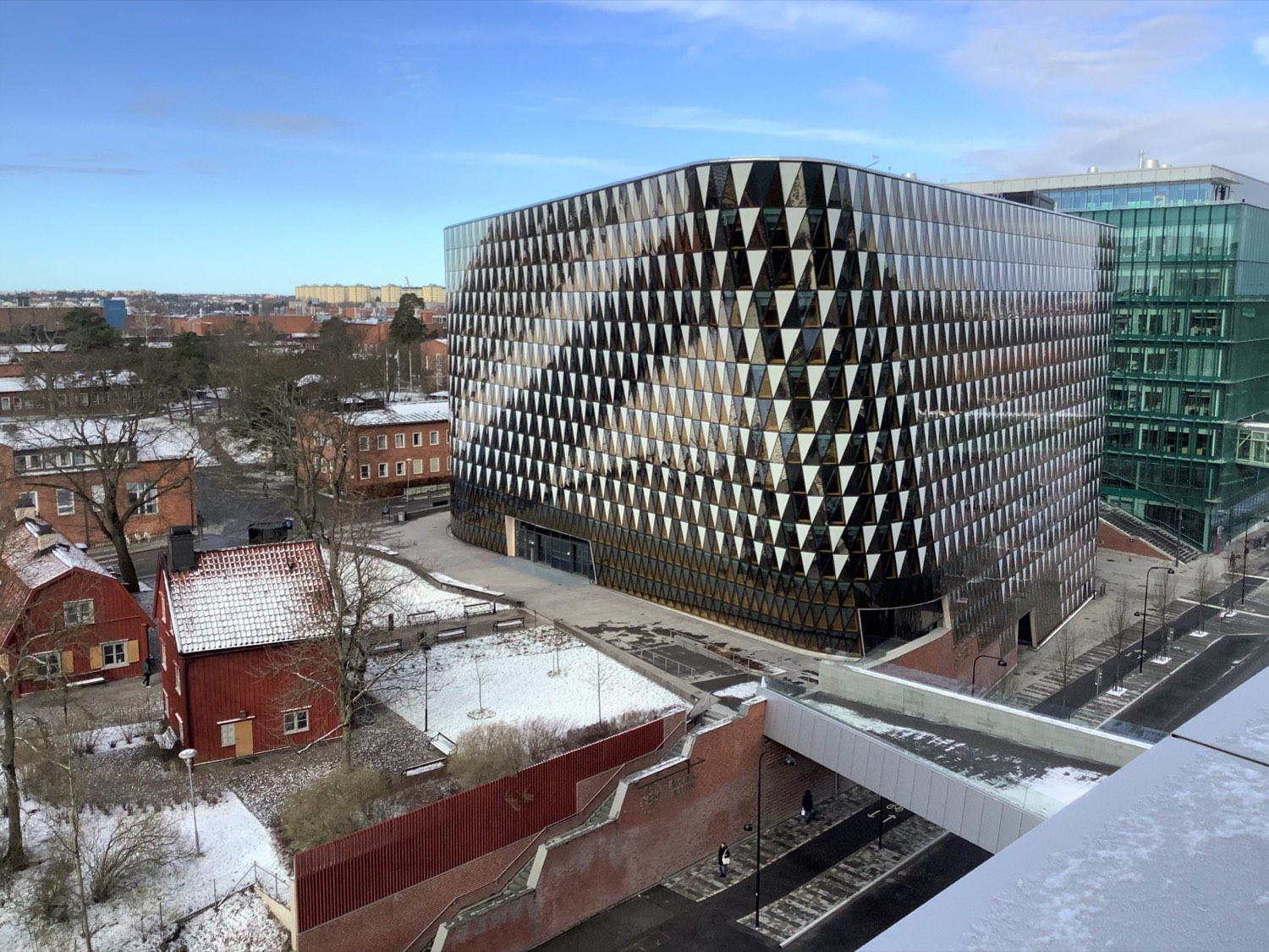 Aula Medica, the Karolinska Institute's striking auditorium building in Solna, Stockholm — a large angular structure clad in a chequered pattern of golden and dark glass panels, set among older red-brick buildings under a blue winter sky.