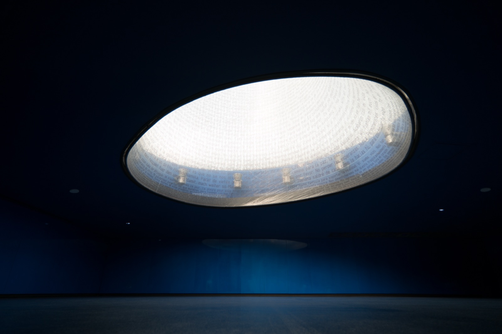 The interior of the 11-M memorial at Atocha station — looking up inside a tall dark chamber toward a glowing oval skylight formed by a translucent membrane inscribed with thousands of messages of condolence, lit from within against the deep blue of the surrounding walls.