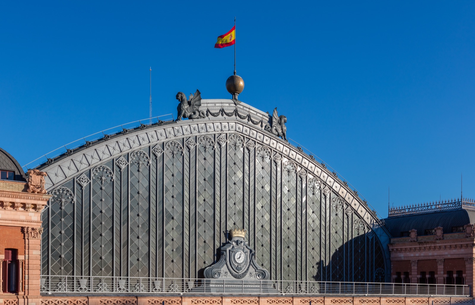 The great glazed train-shed roof of Madrid's Atocha railway station, photographed from outside against a deep blue sky — a vast arched canopy of grey iron and glass topped by sculptures and a Spanish flag, with ornate brick station buildings flanking it.
