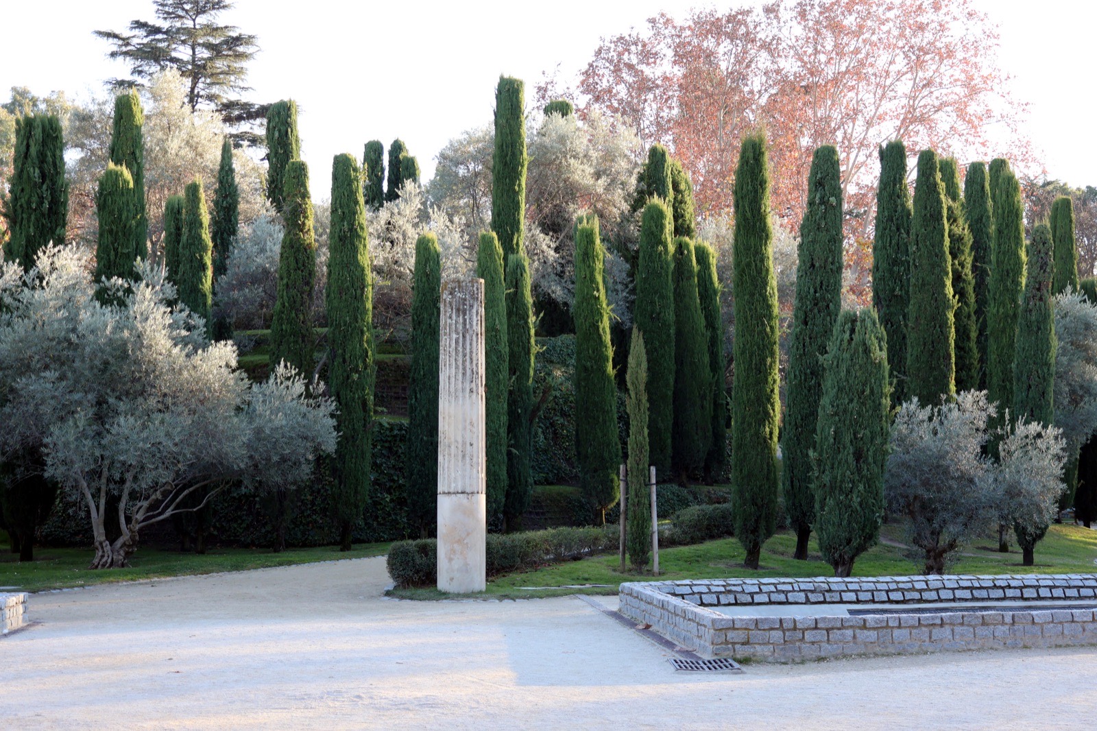 The Bosque del Recuerdo in Madrid's Retiro park — a memorial grove of olive and cypress trees planted on a low mound surrounded by a ring of water, one tree for each person killed in the 2004 bombings, under a clear sky.
