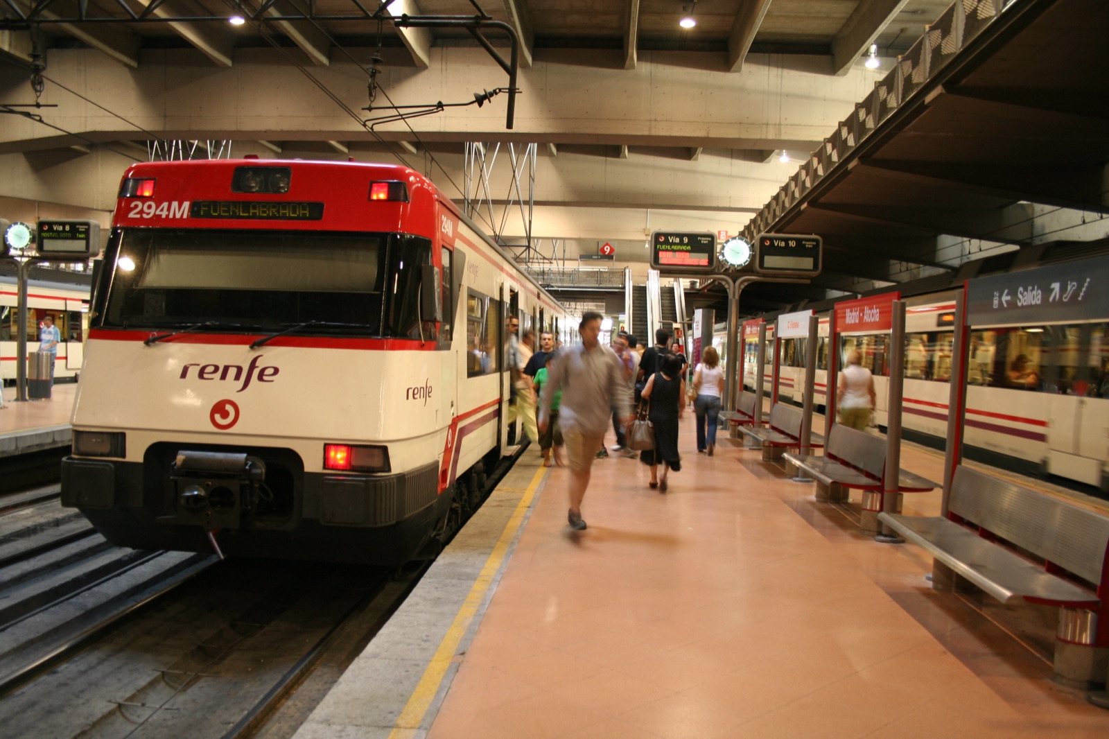 A red-and-white Cercanías commuter train of the Madrid suburban network standing at a platform at Atocha, its double-deck carriages stretching along the platform under the station's canopy.