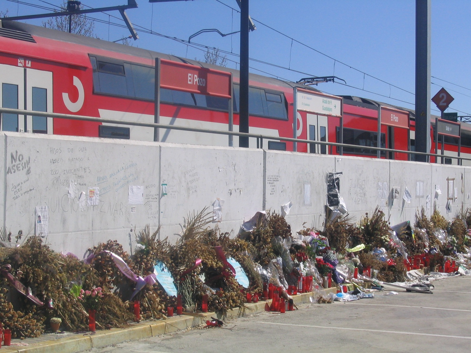 The platform at El Pozo station in 2004, weeks after the bombing — a red Cercanías train marked 'El Pozo' standing on the line, and along the foot of the platform's concrete retaining wall a long spontaneous memorial of hundreds of red candle-jars, wilted flower bouquets, and handwritten notes and graffiti, including the Spanish words 'No al asesinato.'