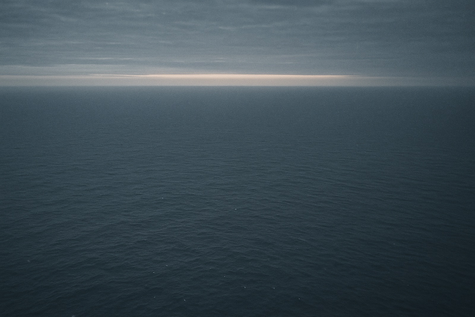A vast empty stretch of the southern Indian Ocean at twilight, photographed from approximately 15,000 feet — horizon-to-horizon open water with small white-caps, no land or vessels visible, thin overcast above and the last band of pale orange sun on the curved horizon.