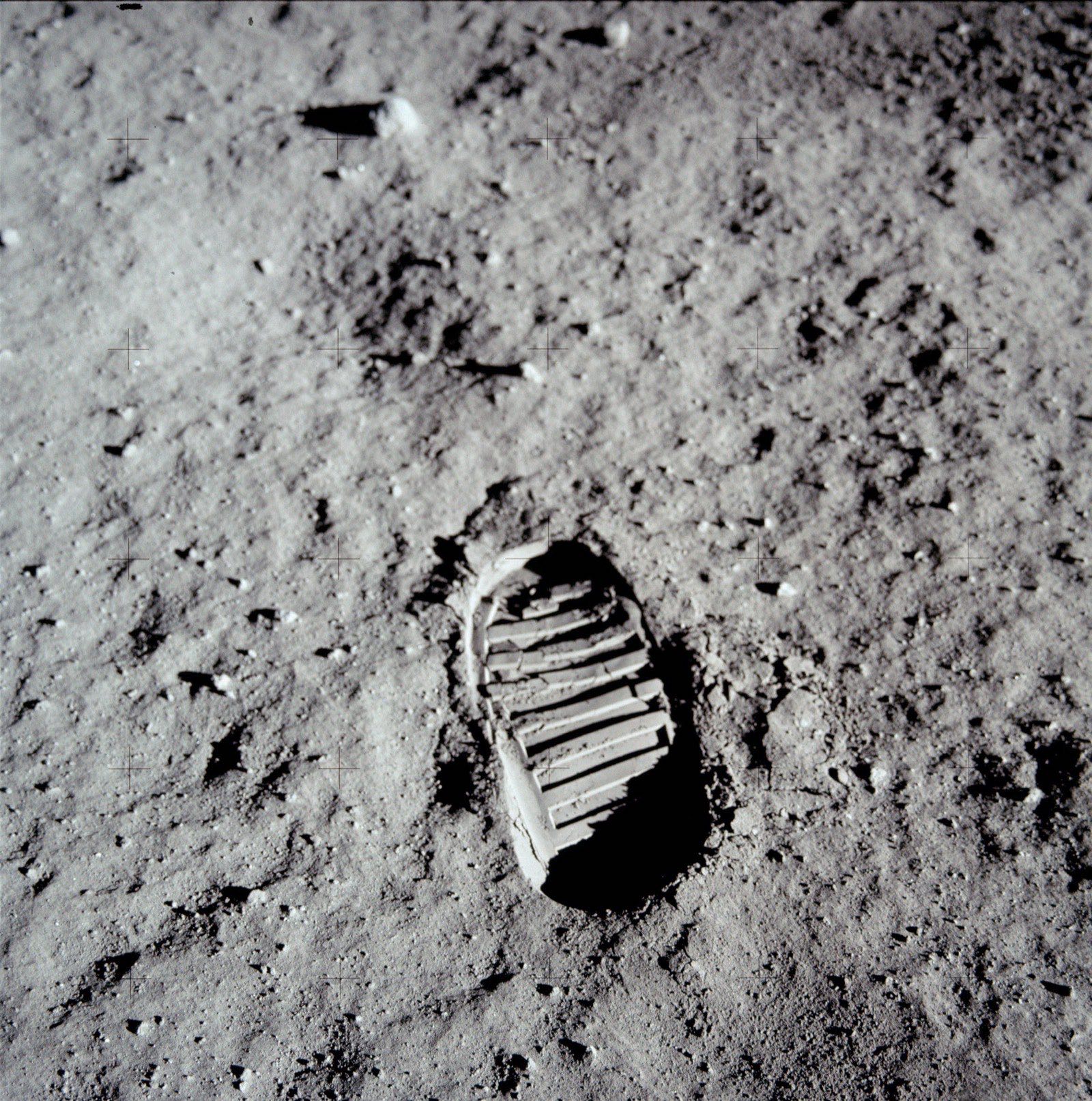 A close-up photograph of Buzz Aldrin's bootprint pressed into the dusty lunar regolith. The print is crisp and detailed, with the bumpy texture of the boot sole clearly visible against the fine grey-black surface dust.