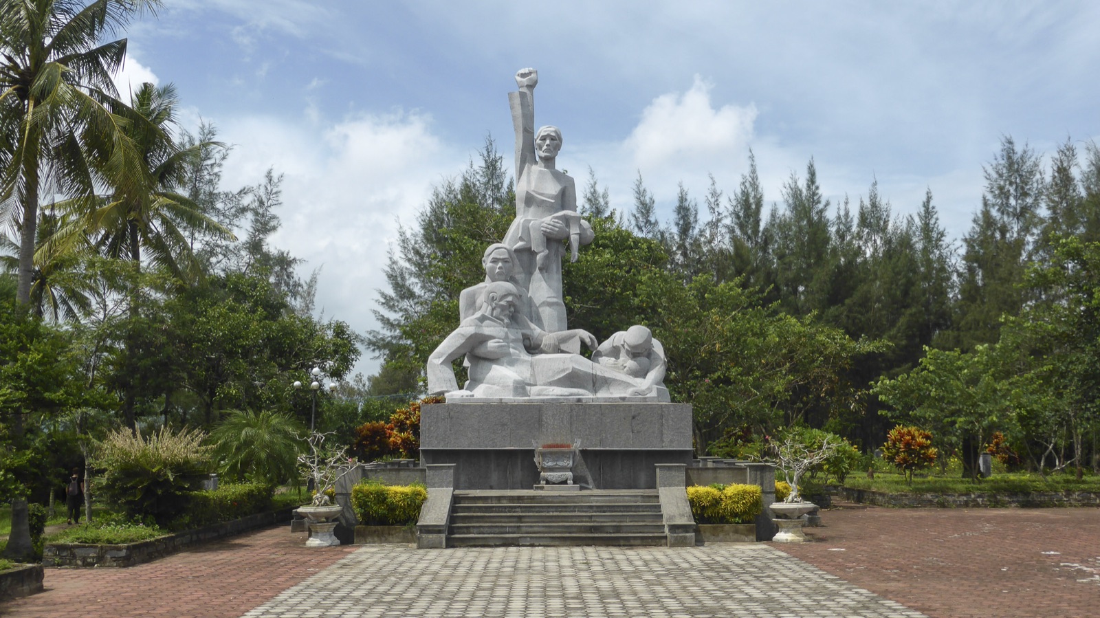 The Sơn Mỹ Memorial Park in Quảng Ngãi Province, Vietnam — a stone monument at the entrance to the memorial complex, surrounded by mature tropical trees and a paved courtyard.