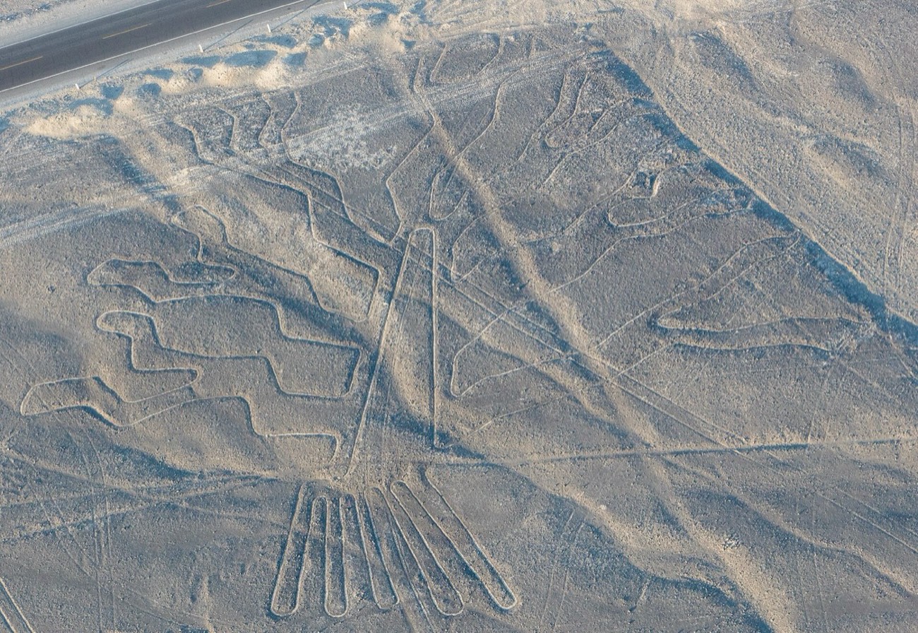 An aerial view of a large Nazca geoglyph on the desert plain, with the Pan-American Highway visible cutting across the top of the frame.