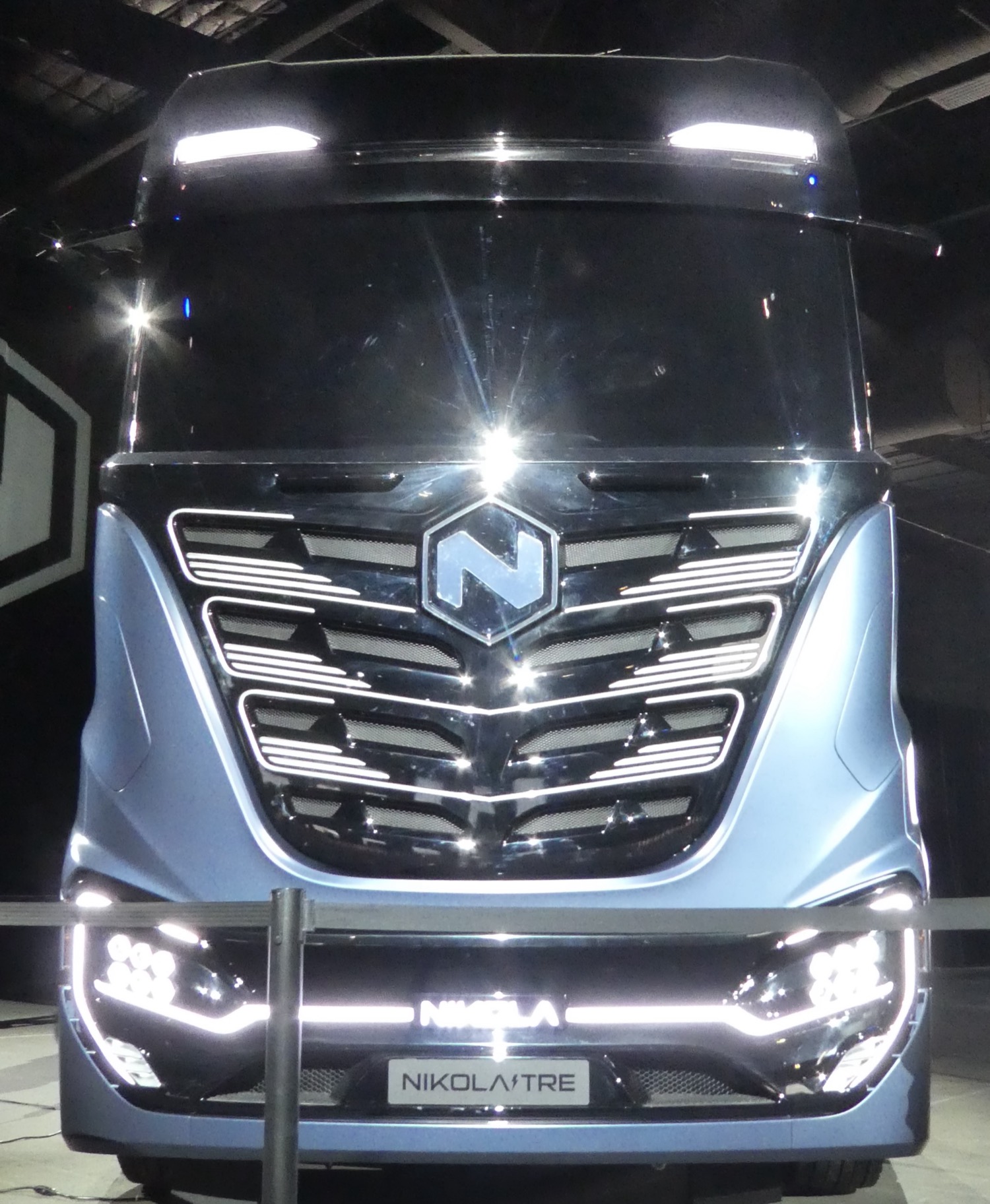 The front of a Nikola Tre electric semi-truck on display, a sleek blue-grey cab with the stylised 'N' Nikola logo on the grille and 'NIKOLA TRE' on a sign beneath it.